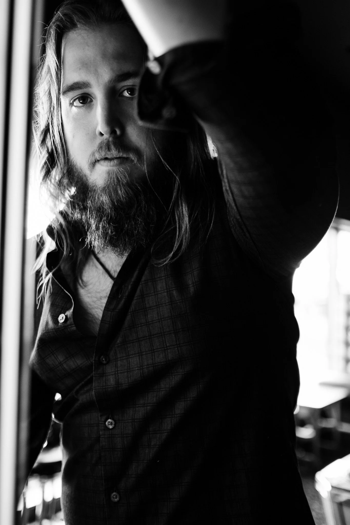 Black and white close-up of a man with long hair and beard adjusting a mirror or window, looking into the camera.