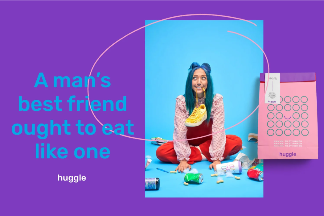 A woman with blue hair sitting on the floor surrounded by snack wrappers and soda cans, holding a sneaker in her mouth, with a pink shopping bag nearby. The background is a split of purple and blue, with text reading "A man’s best friend ought to eat like one" and the brand name "huggle."