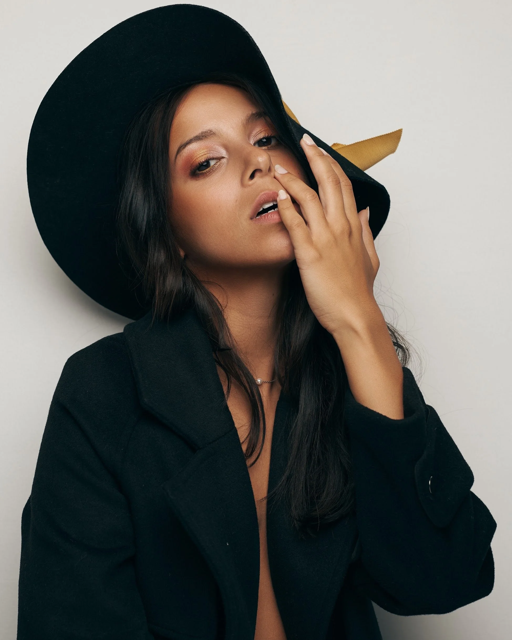 A woman with long dark hair wearing a black wide-brim hat and black coat, posing with her hand near her face against a light background.
