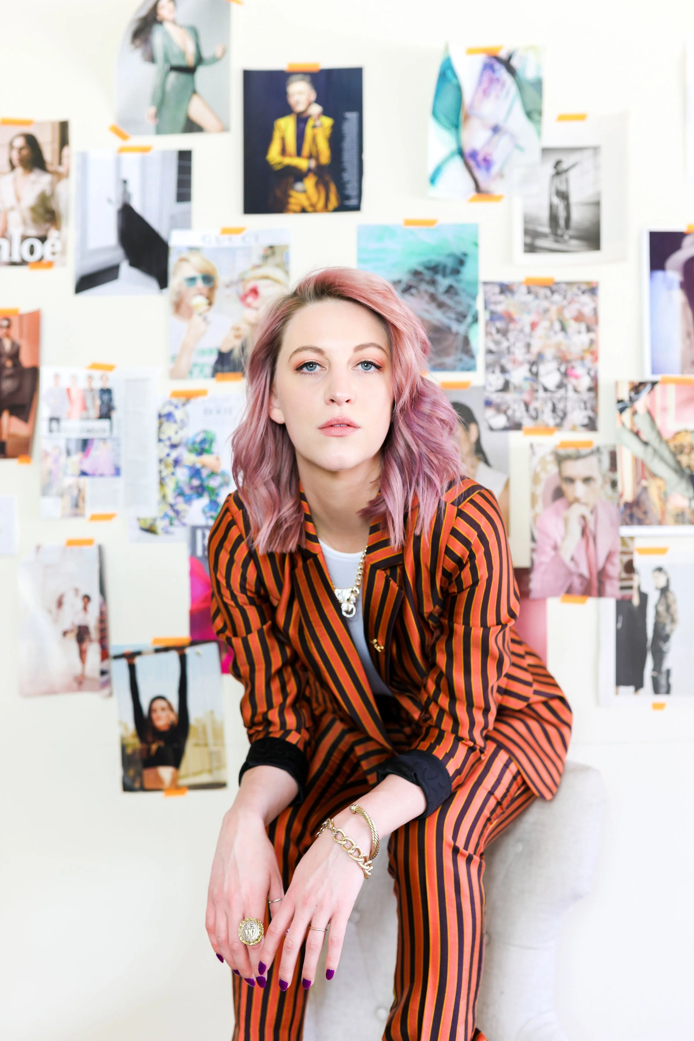 Stylish woman with pink hair in a striped orange and black suit, sitting on a stool in front of a wall covered with fashion photographs and magazine clippings.