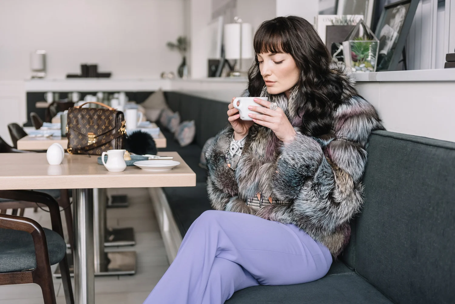 A woman with dark hair in a fur coat from ML Furs sitting in a cafe, drinking from a white mug, with a Louis Vuitton bag and tableware on the table.