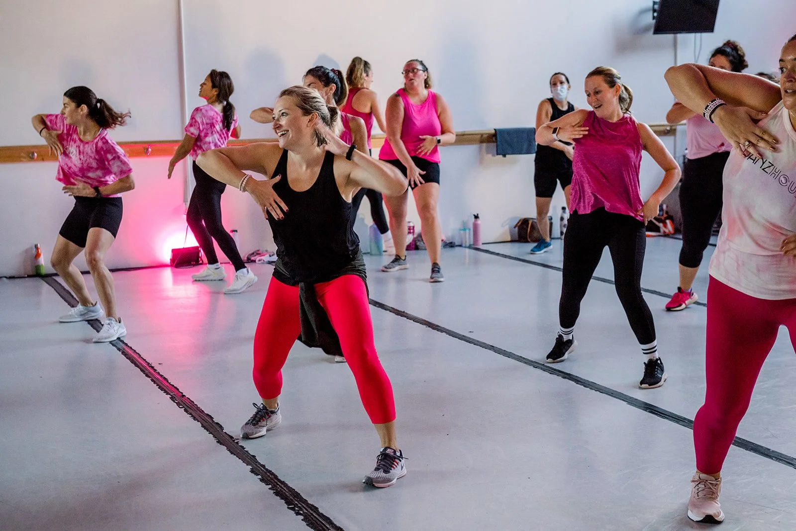 Group of women participating in a dance fitness class in a studio, wearing workout clothes, some smiling, with a ballet barre and a TV screen on the wall.