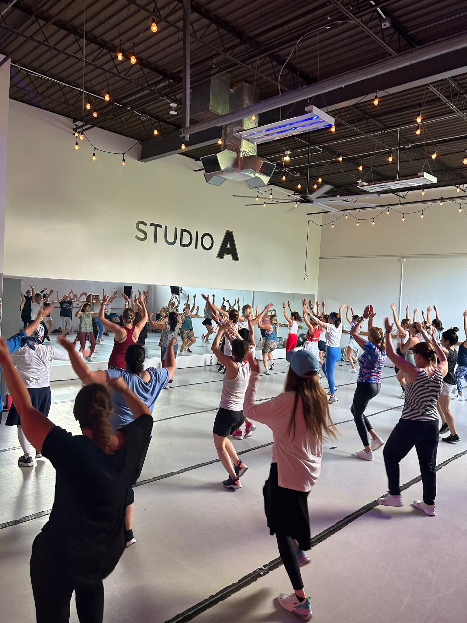 A large group of people participating in a dance or fitness class in a studio labeled 'STUDIO A', with a mirrored wall and ceiling string lights.
