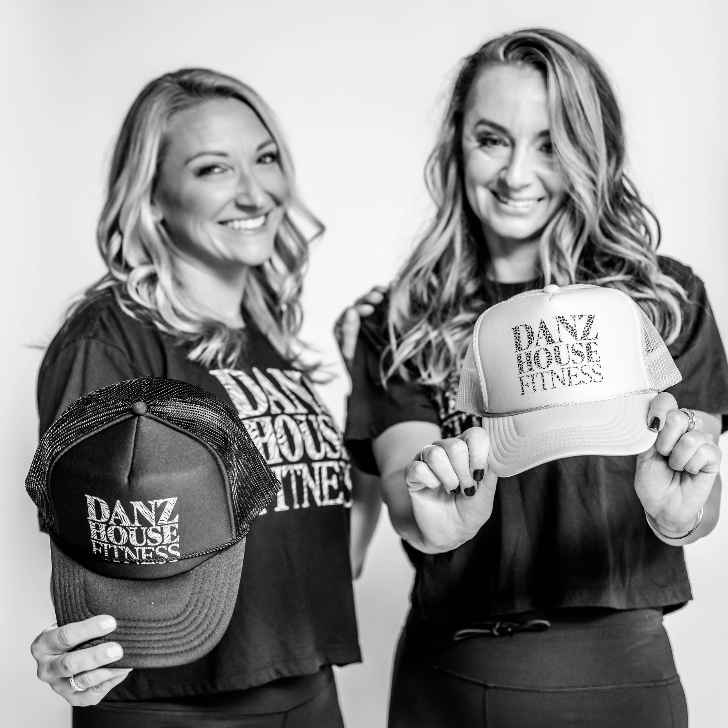 Two women with long wavy hair smiling, holding hats with 'DANZ HOUSE FITNESS' logos, wearing black T-shirts, in front of a plain light-colored background.