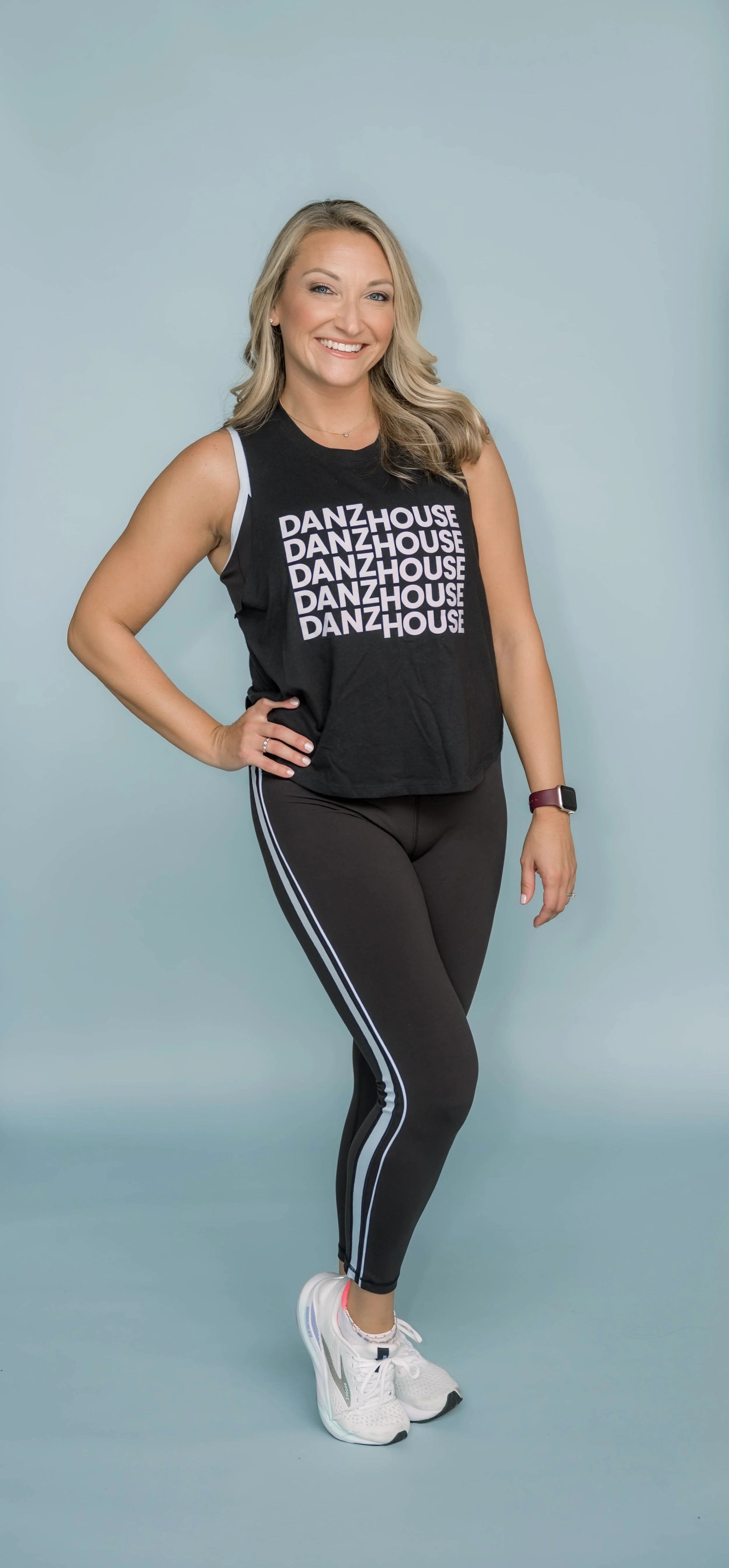 A woman in athletic wear, including a black tank top with the word 'DANZHOUSE' repeated in white, black leggings with white and blue stripes on the sides, and white athletic shoes, standing against a plain light blue background, smiling with one hand on her hip.