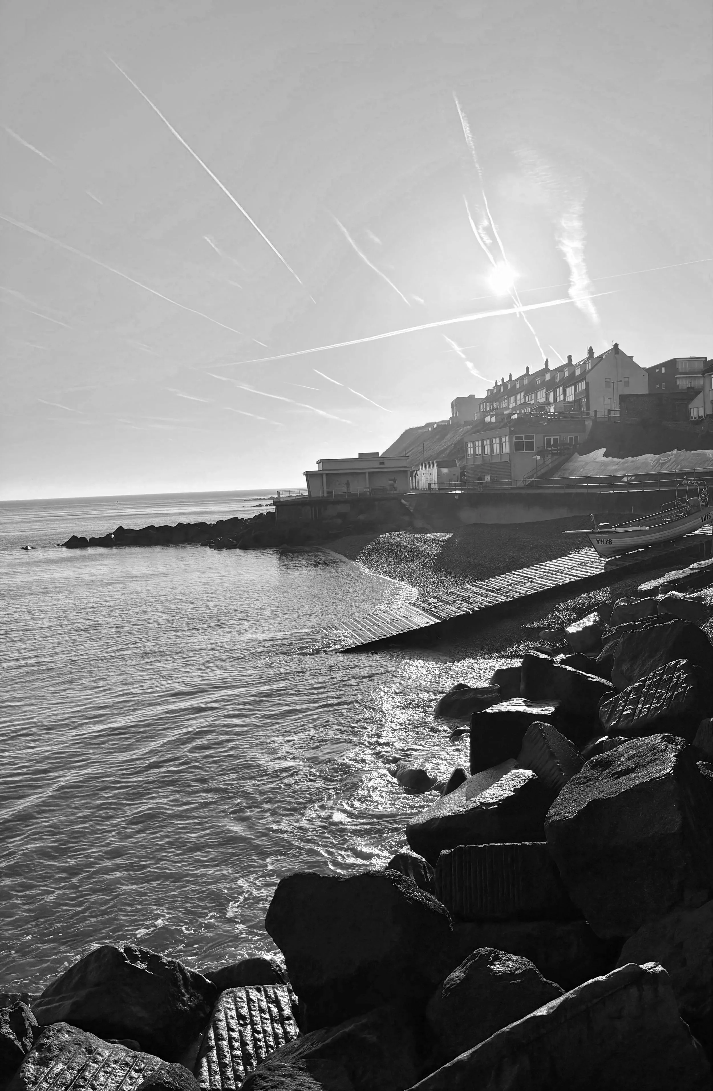 A black and white photo showing a coastal scene with large rocks along the shoreline, a small boat on the water, and houses perched on a hillside under a sky with streaks from contrails.