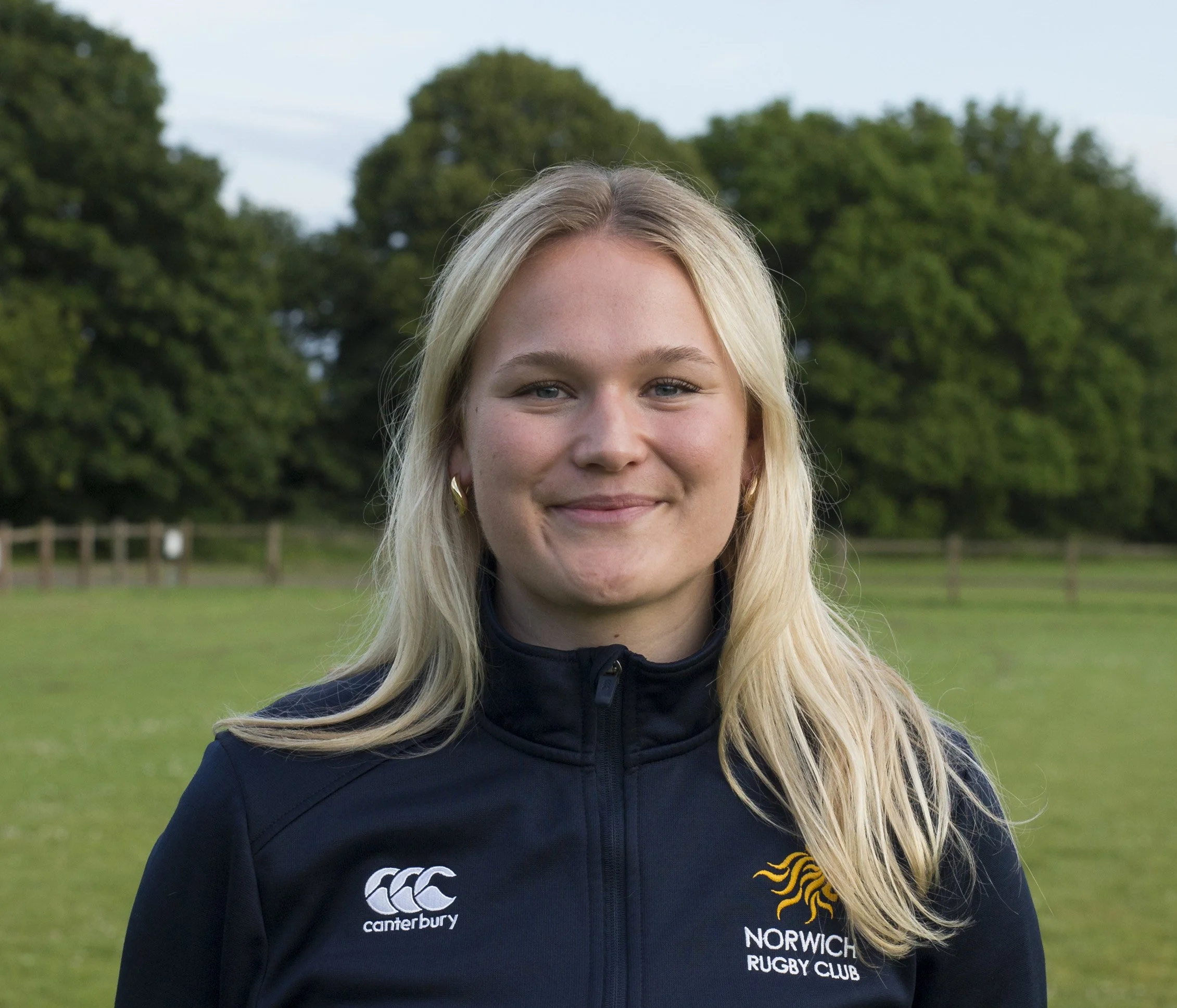 A young blonde woman smiling outdoors in a rugby jacket with Norwich Rugby Club logo, standing in a grassy field with green trees in the background.