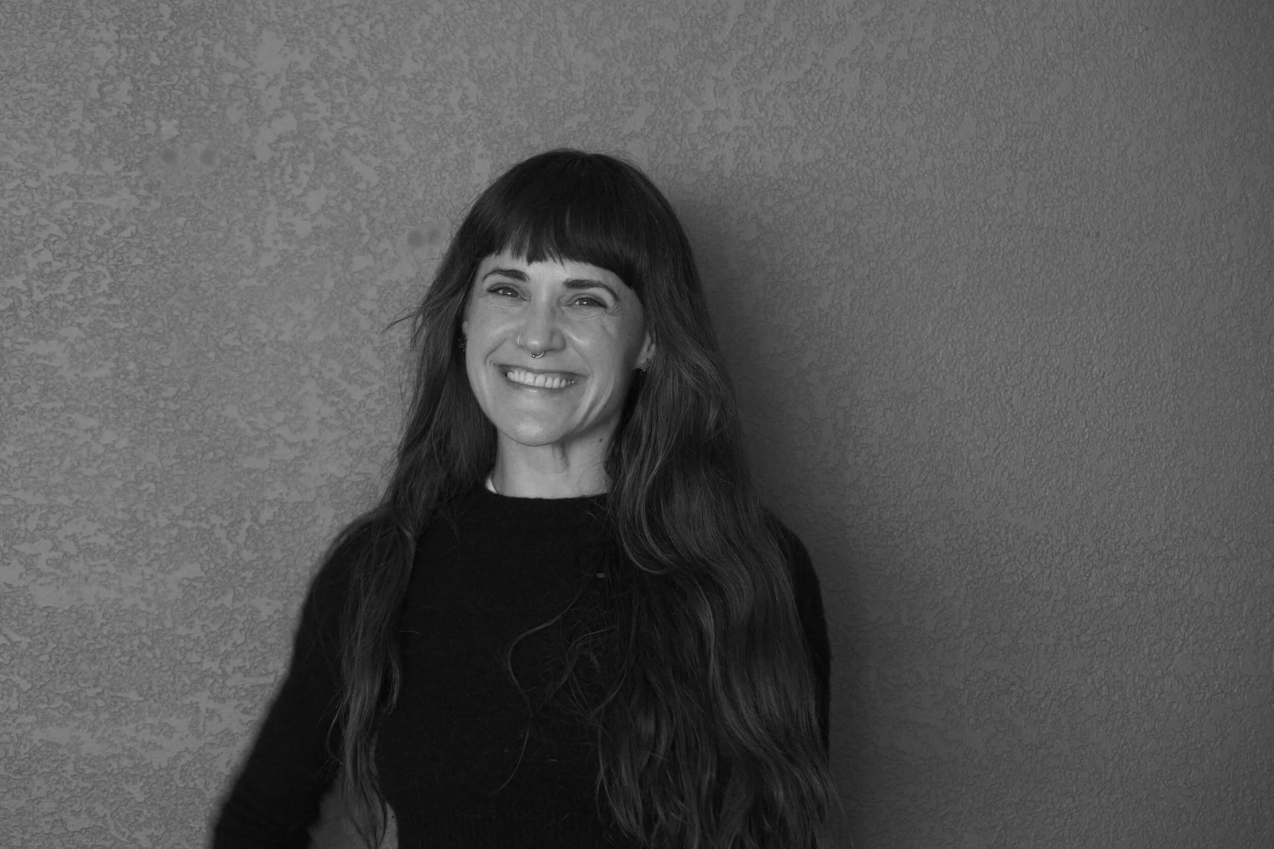 A woman with long, wavy hair and a nose ring, standing against a textured wall, smiling in black and white.