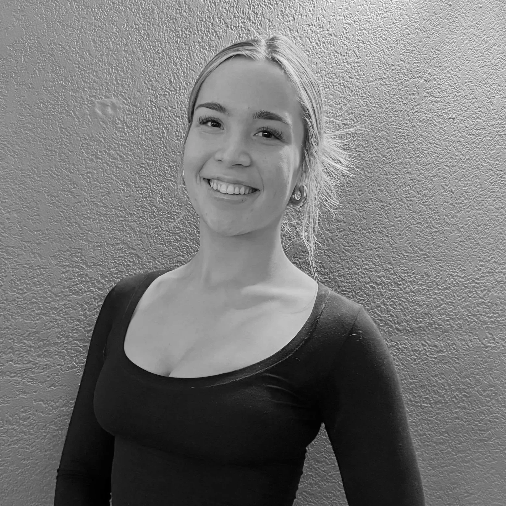 A young woman with blonde hair smiling, wearing a black long-sleeve top, standing in front of a textured wall.