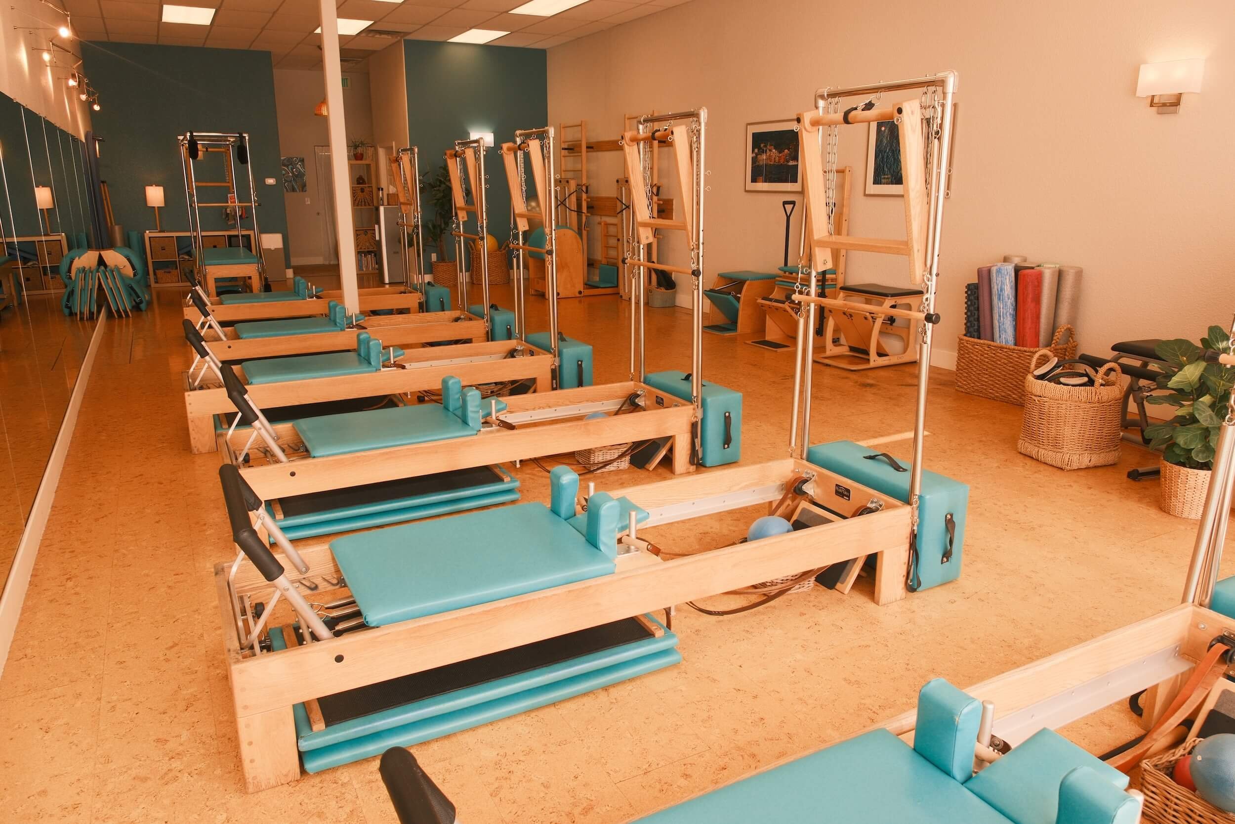Up to six Pilates reformer machines with teal pads and metal frames arranged in a row in a well-lit studio