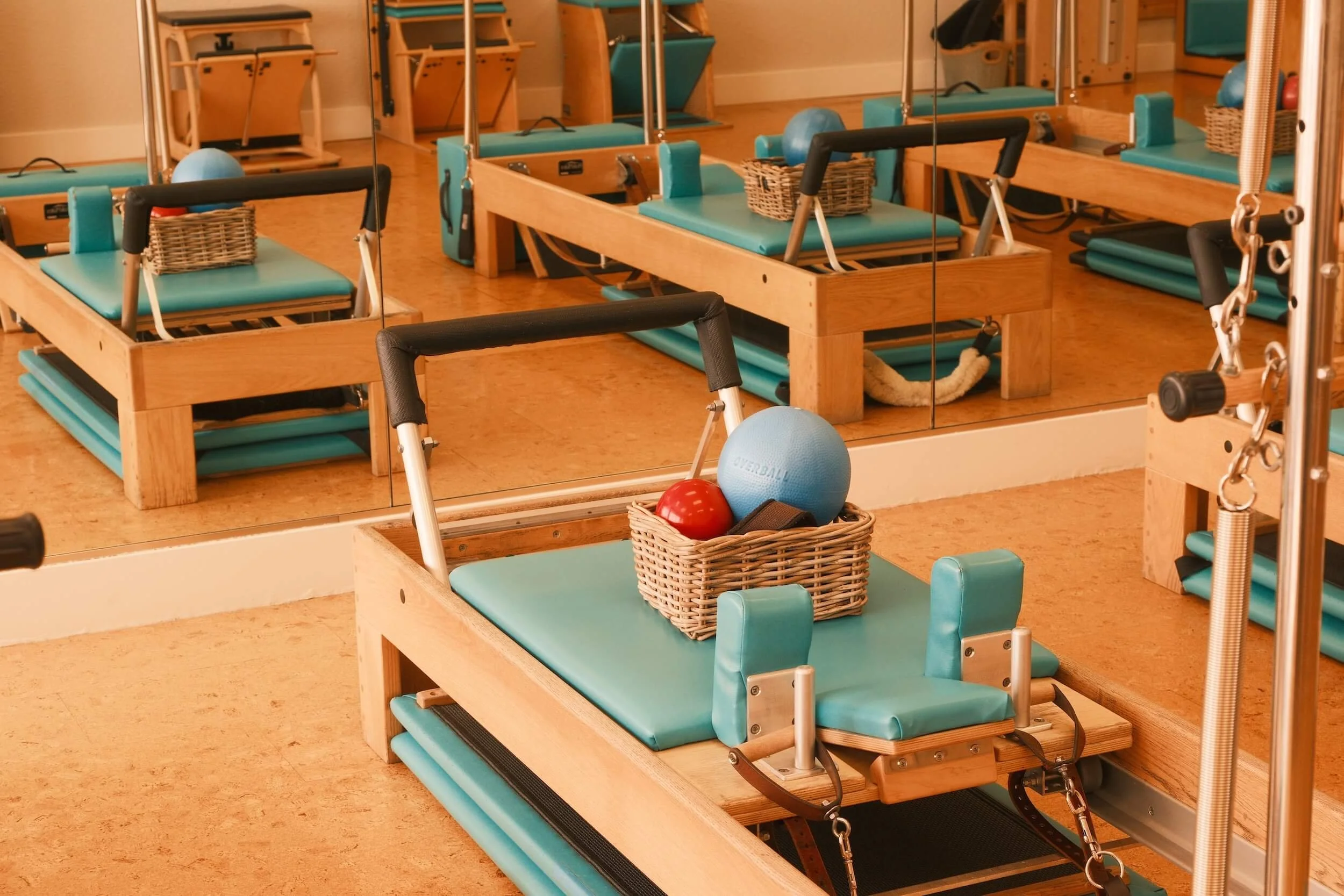 Pilates reformer machines with turquoise padding and wooden frames, some with baskets holding exercise balls and small weights, in a fitness studio.
