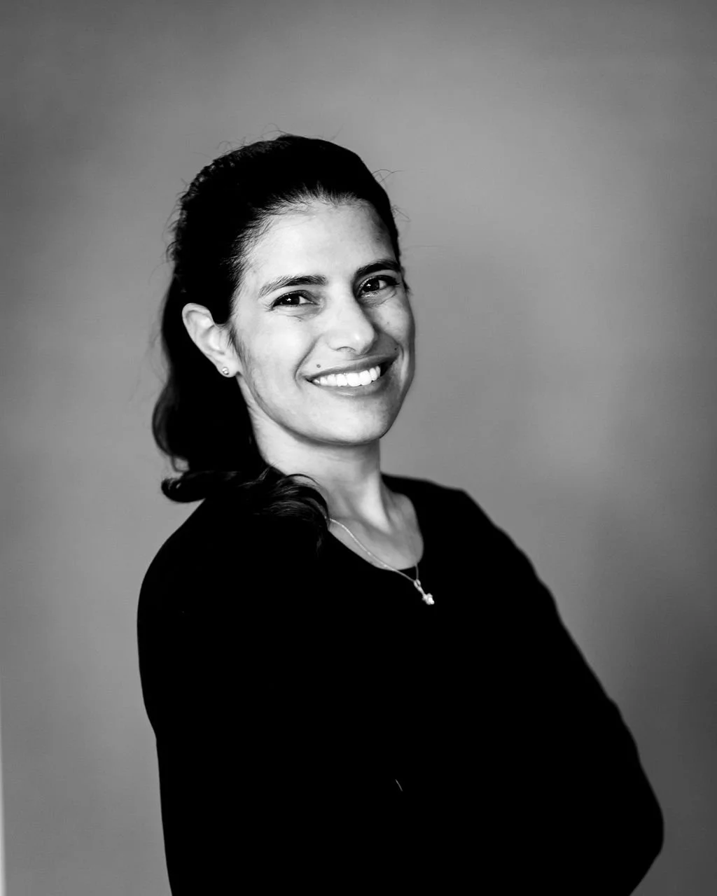 Black and white portrait of a woman with dark hair tied back, smiling, wearing a dark top and a necklace, against a plain background.