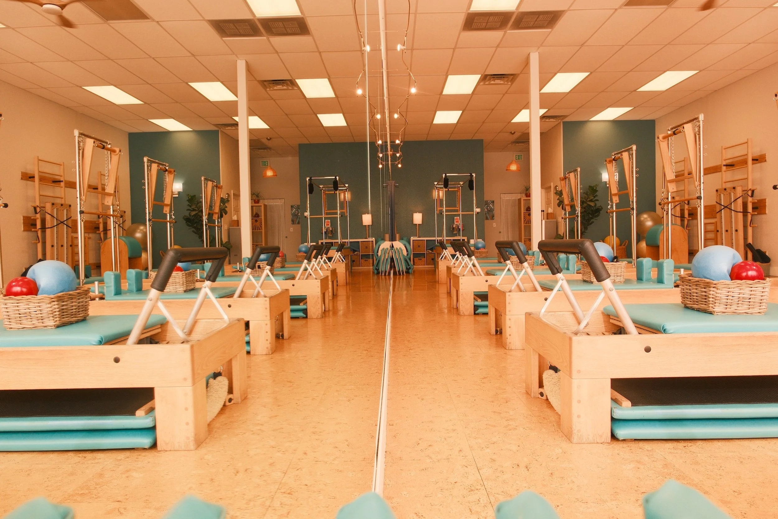 An indoor Pilates studio with mirrored wall, wooden furniture, exercise mats, stability balls, and Pilates reformer machines arranged in rows.