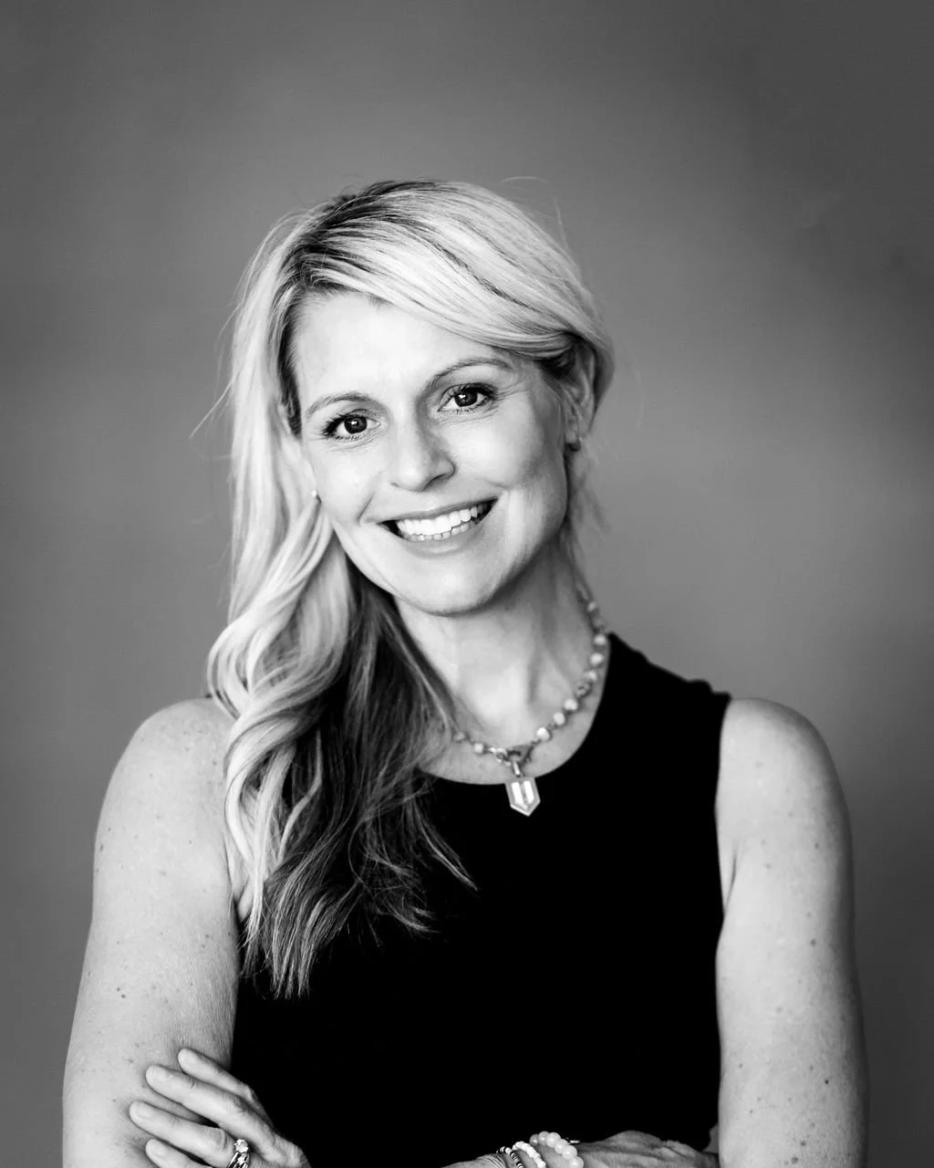 Black and white portrait of a smiling woman with wavy blonde hair wearing a sleeveless black top, a beaded necklace, and a ring on her finger, against a plain background.