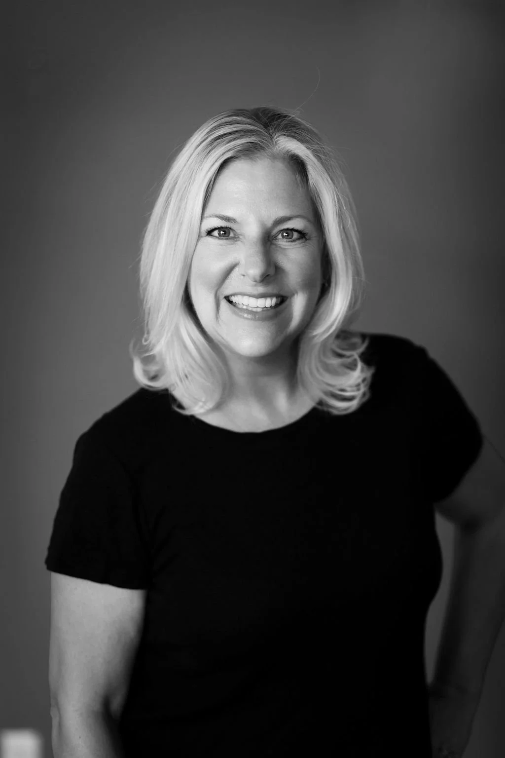 Black and white portrait of a woman with blonde hair smiling, wearing a black top, shot against a plain background.