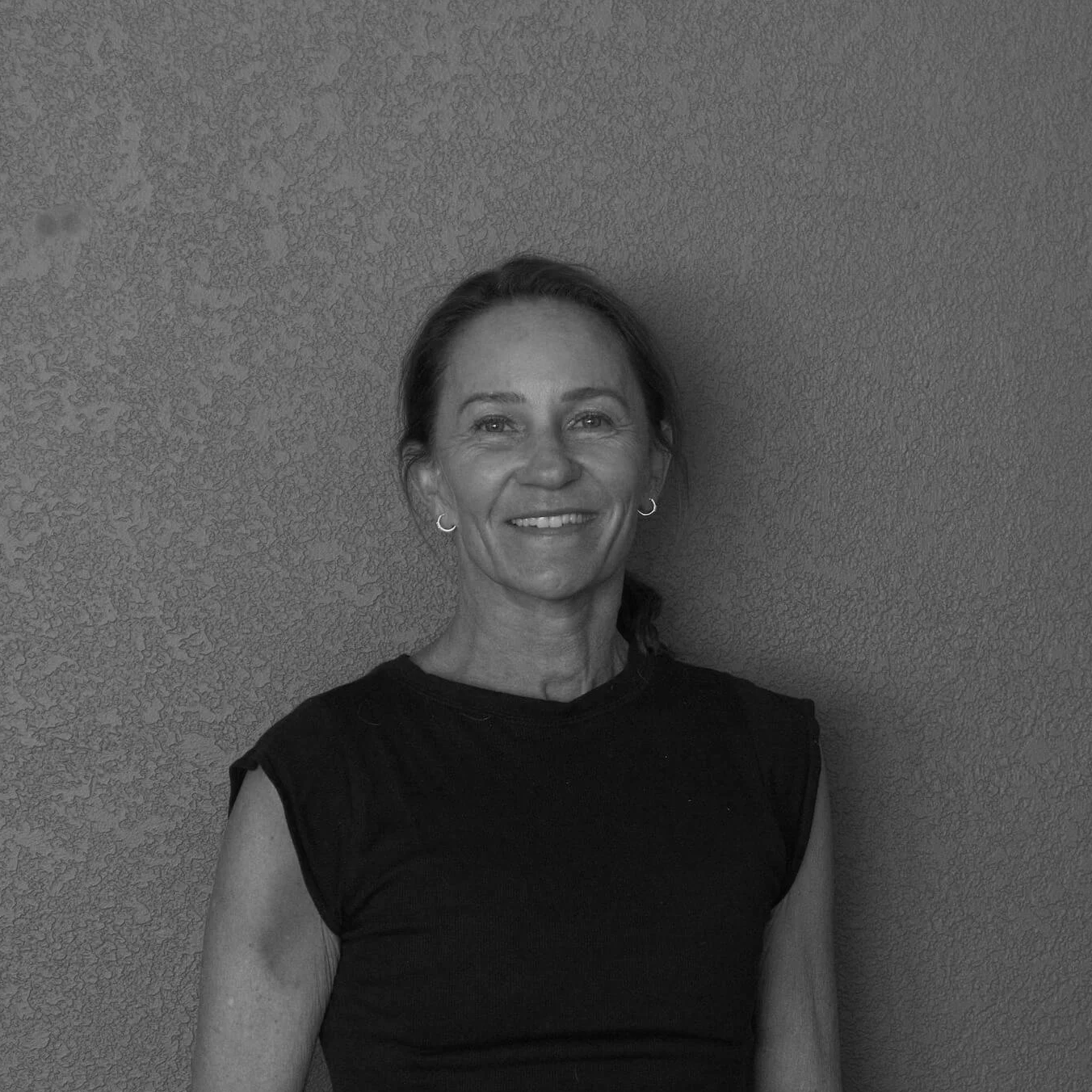 A smiling woman with shoulder-length hair wearing earrings and a sleeveless black top, standing against a textured wall.