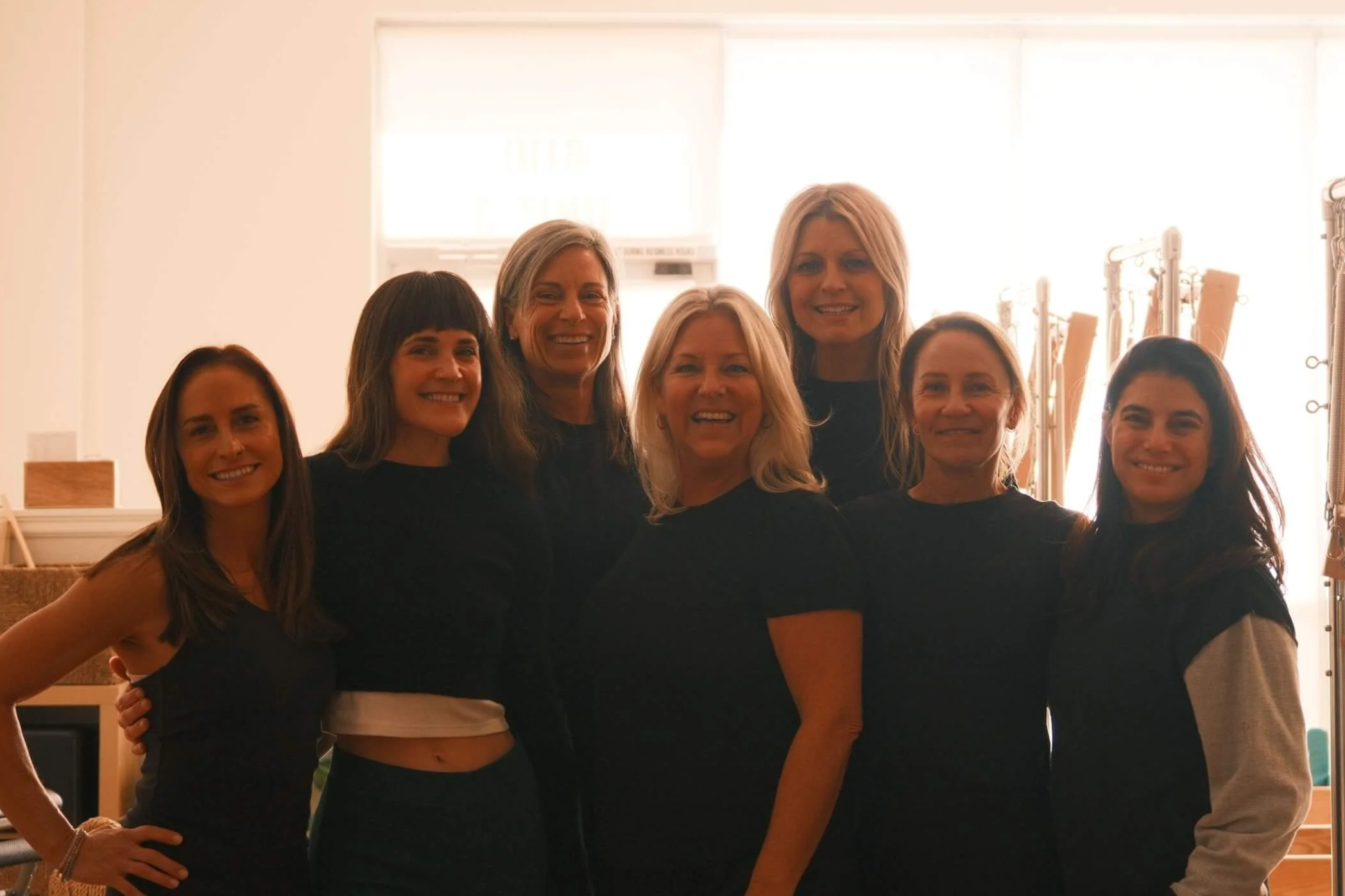 Seven women smiling and posing together indoors with natural light coming from behind them.