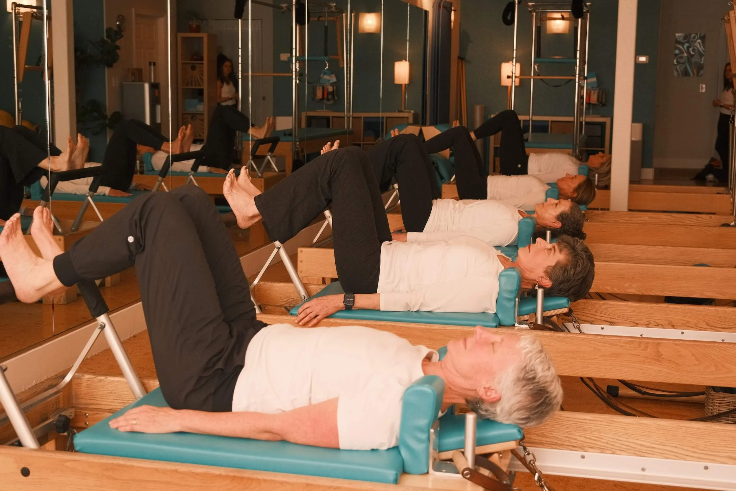 Group of elderly women lying on Pilates reformer beds during a fitness class in a studio with mirrors and wooden decor.