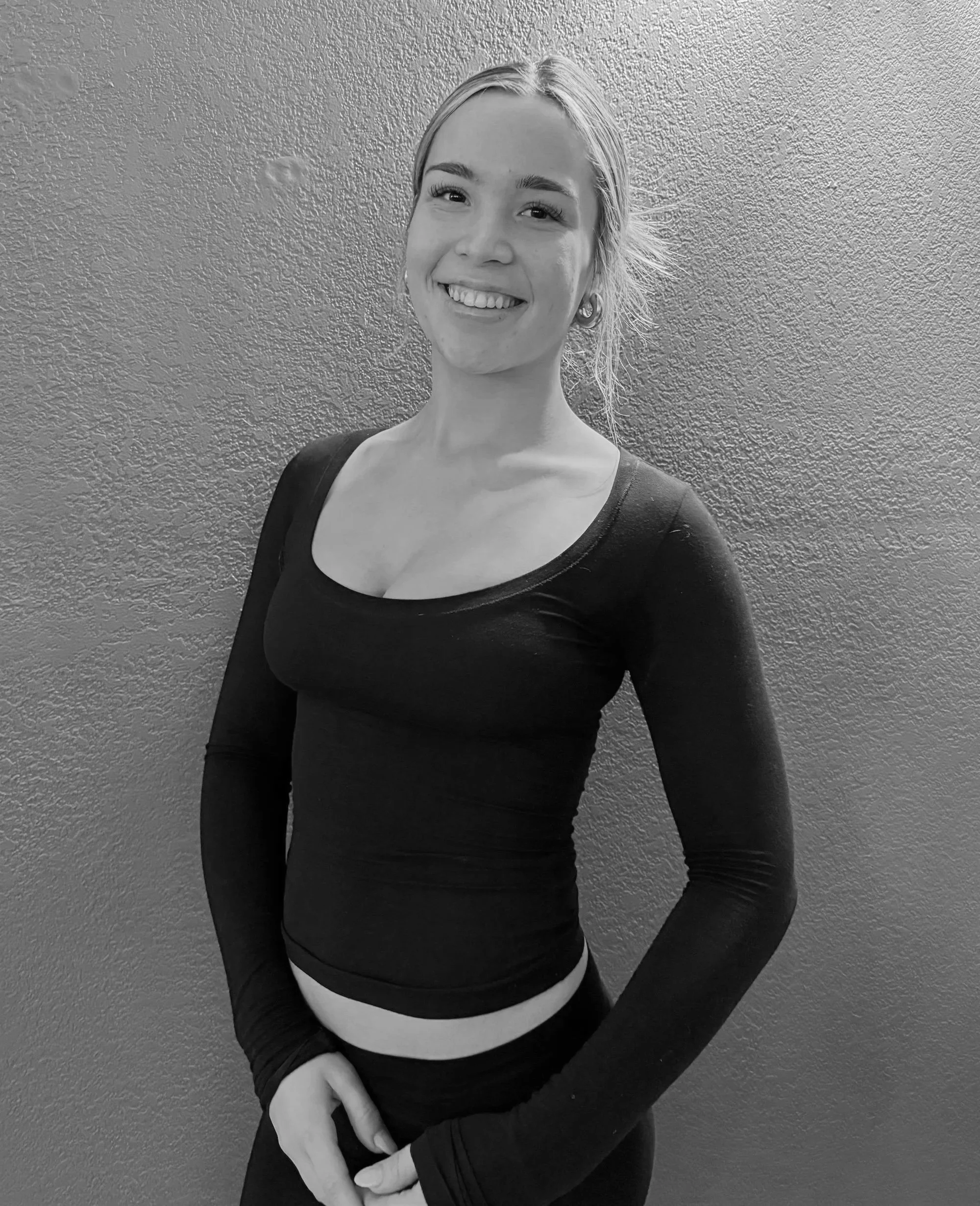 A young woman with a ponytail, smiling, wearing a long-sleeved black top and black pants, standing against a textured wall.