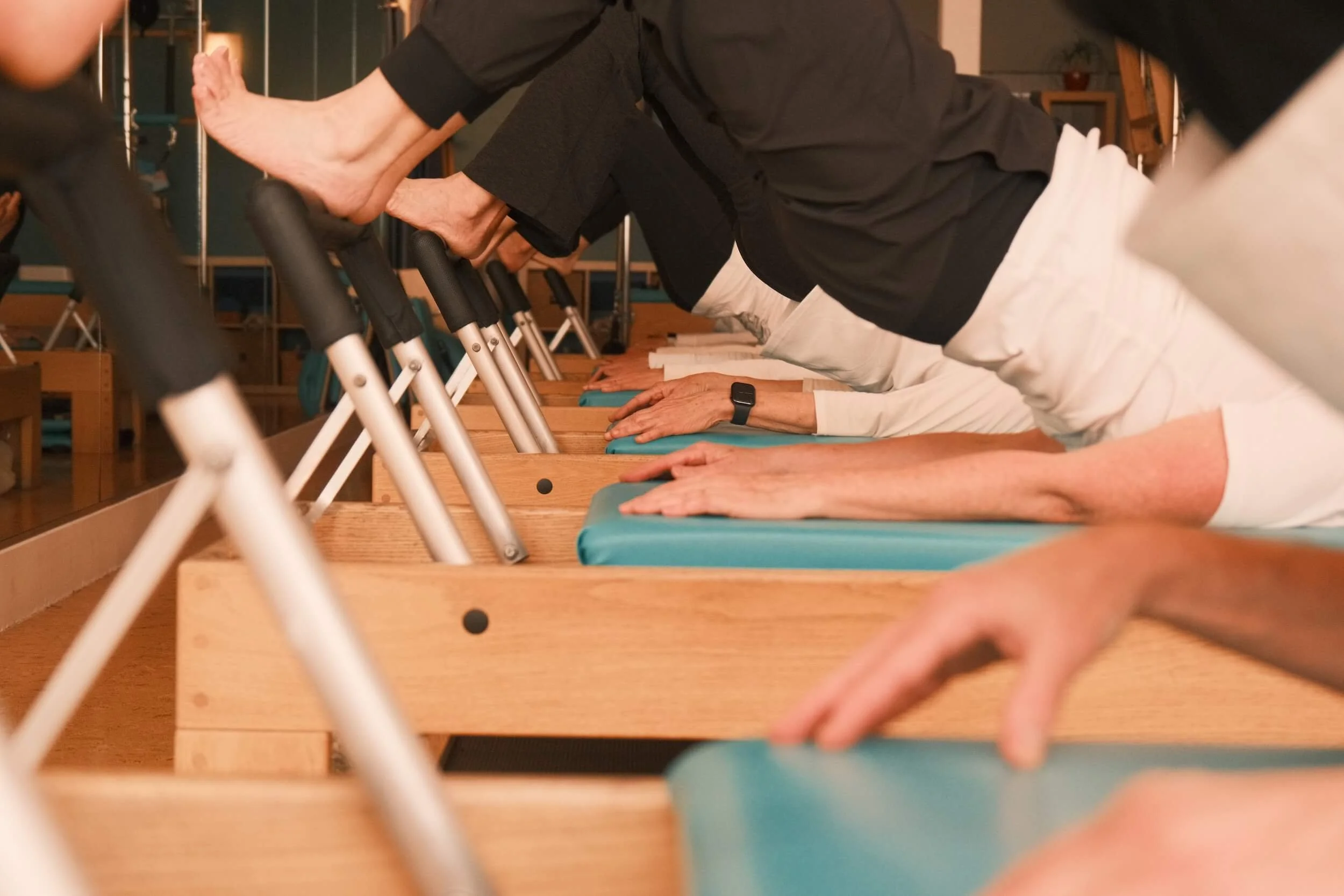 People participating in a yoga class, doing a plank pose on mats, with their hands on the floor and bodies flat, in a studio with wooden floors and a mirror.
