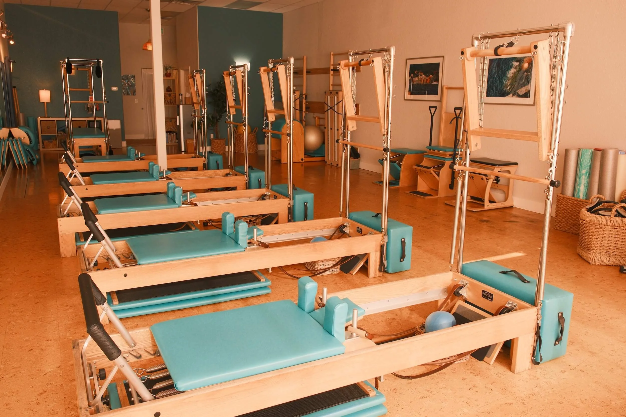 A room with multiple Pilates reformer machines arranged in rows, with teal cushions and wooden frames, set up in a wellness or fitness studio.