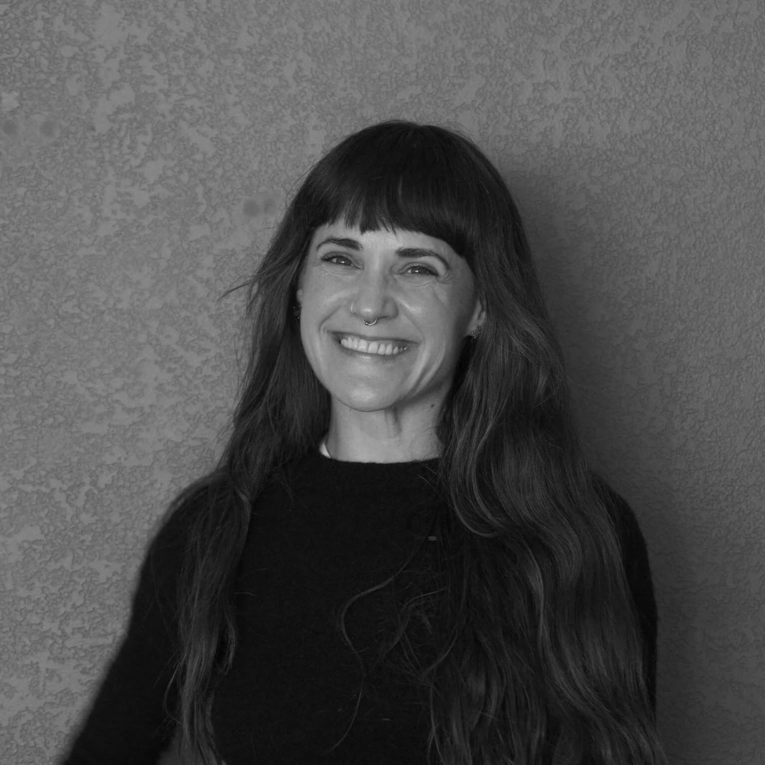 A woman with long wavy hair and bangs, smiling in front of a textured wall.