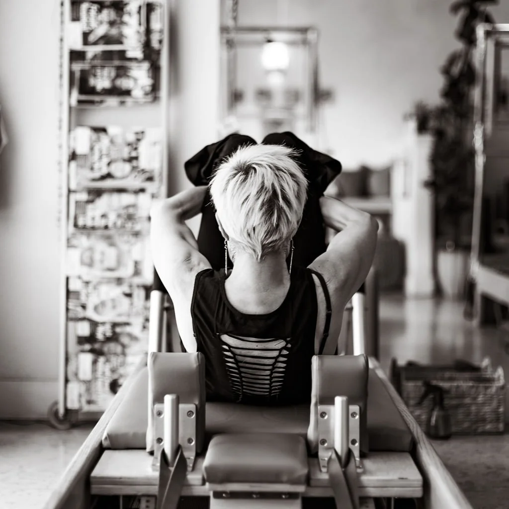 A woman with short blonde hair sitting in a dental chair with her arms raised, holding a black jacket behind her head in a dental office.