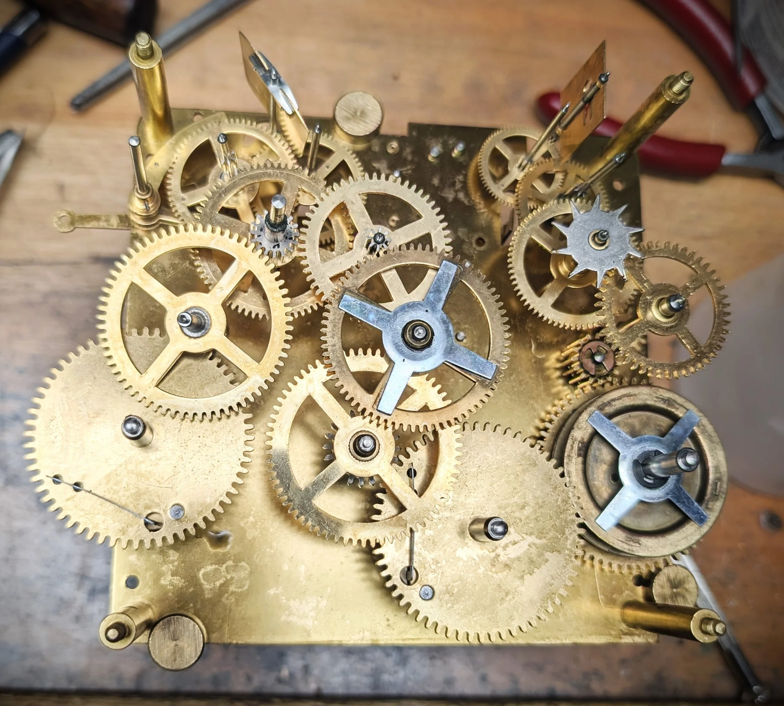Close-up of the intricate gears and mechanisms inside a clock movement, showing brass and steel components in a wooden workspace.