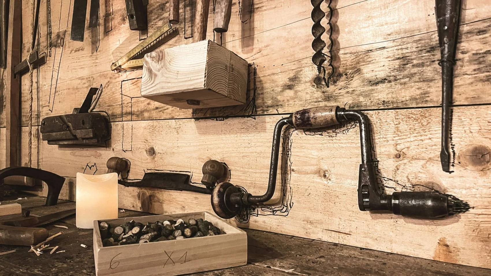 A wooden wall with various old tools mounted, including a hacksaw, an auger bit, and a pipe wrench. Below, there is a wooden surface with a box of nails and other miscellaneous tools, and a candle lit nearby.