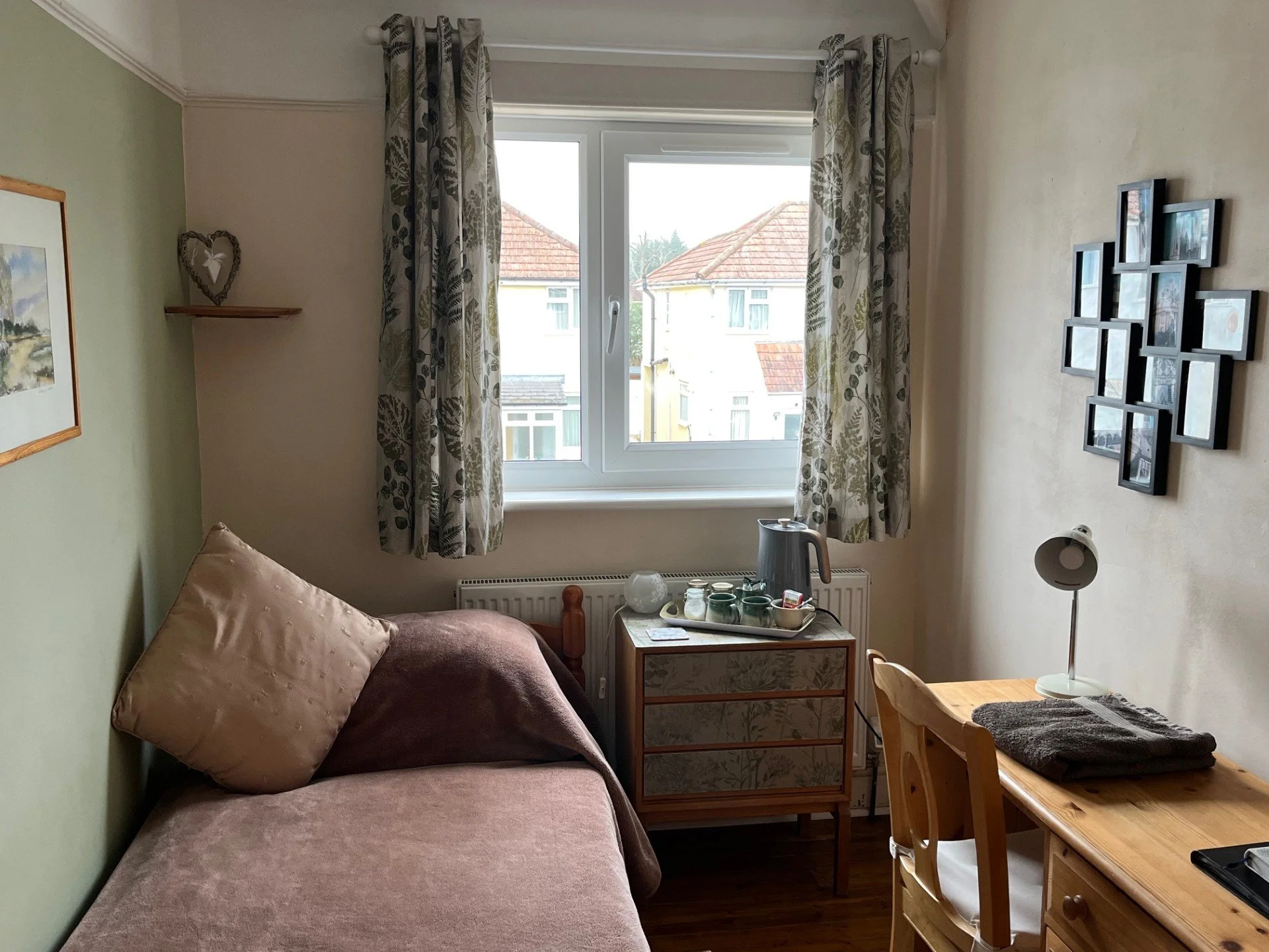 A cozy bedroom with a pink bed, a wooden desk with folded towels and a lamp, a small dresser with a tea set, and a window with floral curtains overlooking neighboring houses.