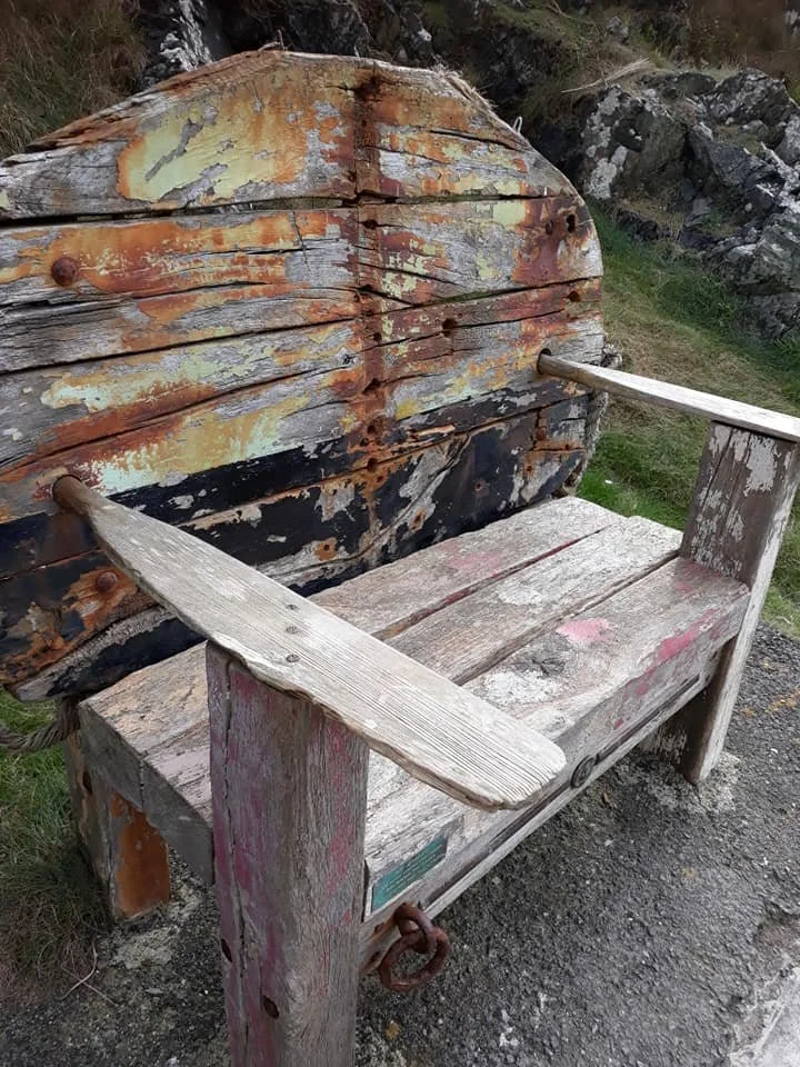 A weathered wooden bench with attached backrest in front of a large, rusted metal pipe.
