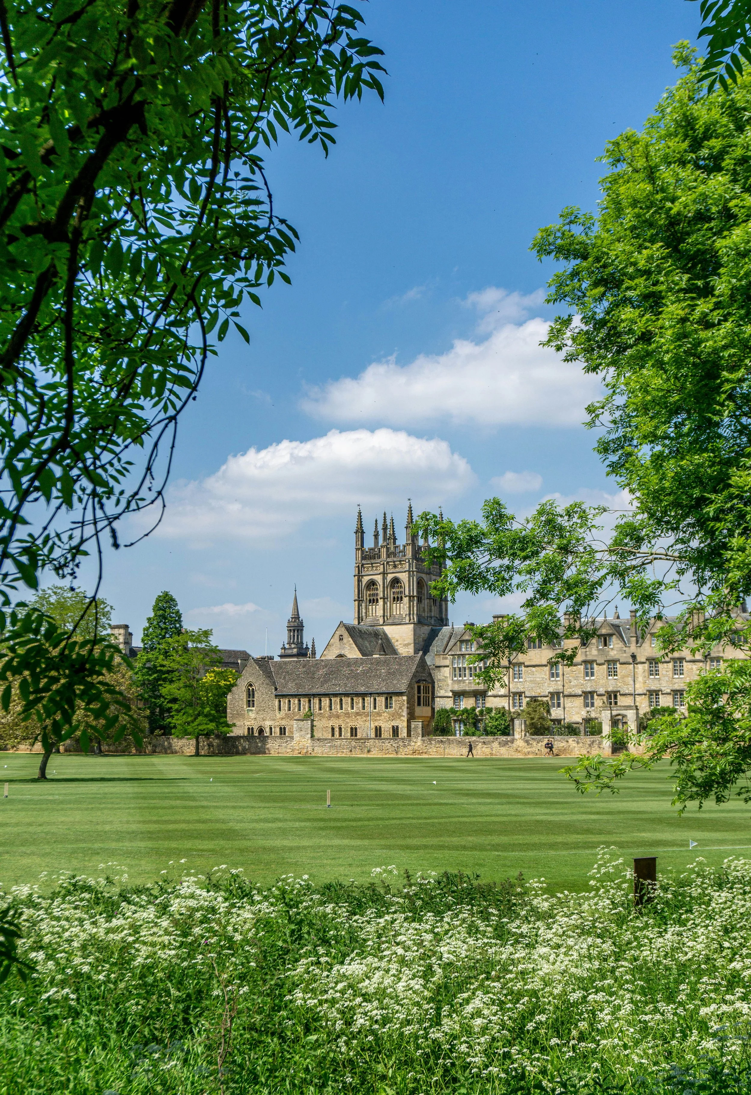 A historic stone church with gothic architecture, surrounded by green trees and a grassy field on a bright, sunny day with some clouds in the sky.