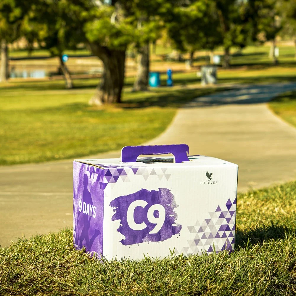 A white and purple box labeled 'C9' and '5 DAYS' resting on grass in a park with trees and a pathway in the background.