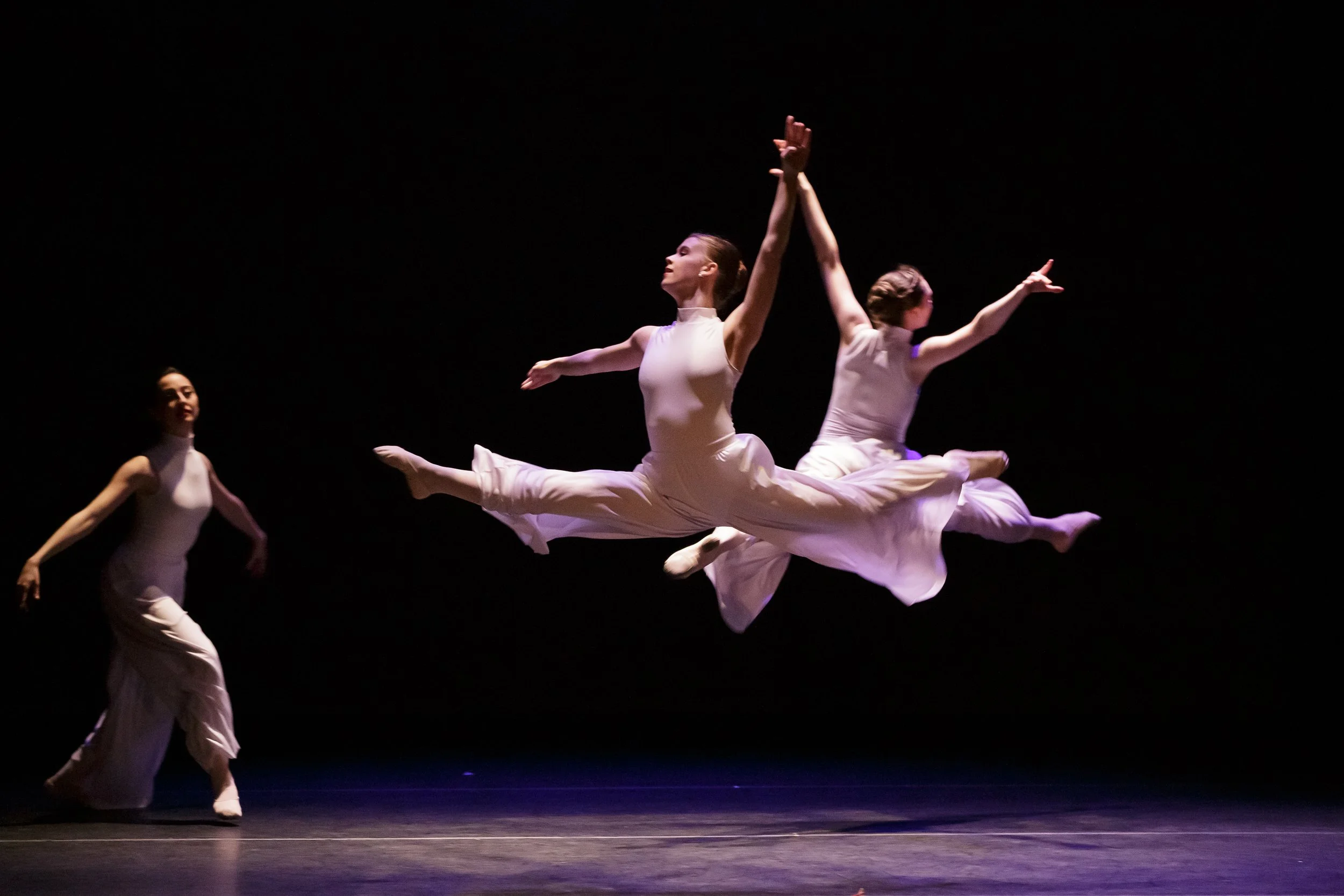 Three ballet dancers performing on stage against a dark background; one dancer is standing on the left, two others are jumping in the air with arms outstretched.