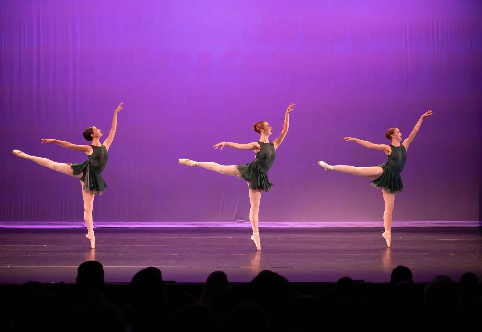 Three ballet dancers in black dresses and tights performing a synchronized dance on stage with purple lighting.