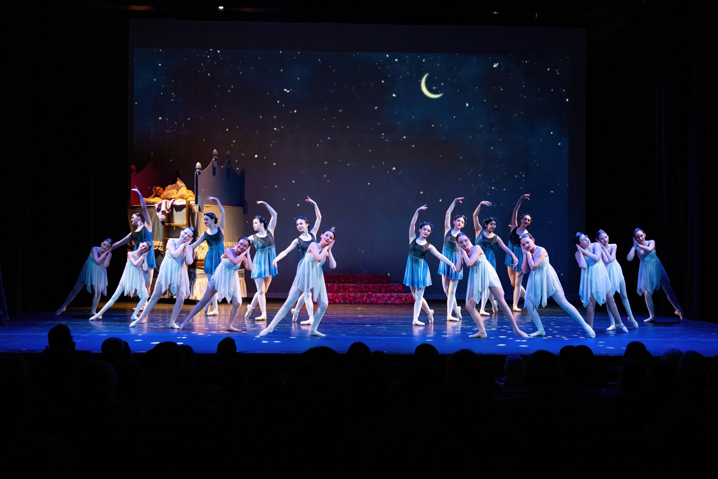 Ballet dancers perform on stage with a night sky backdrop featuring stars and a crescent moon, with some dancers standing and others leaning or arching their bodies.