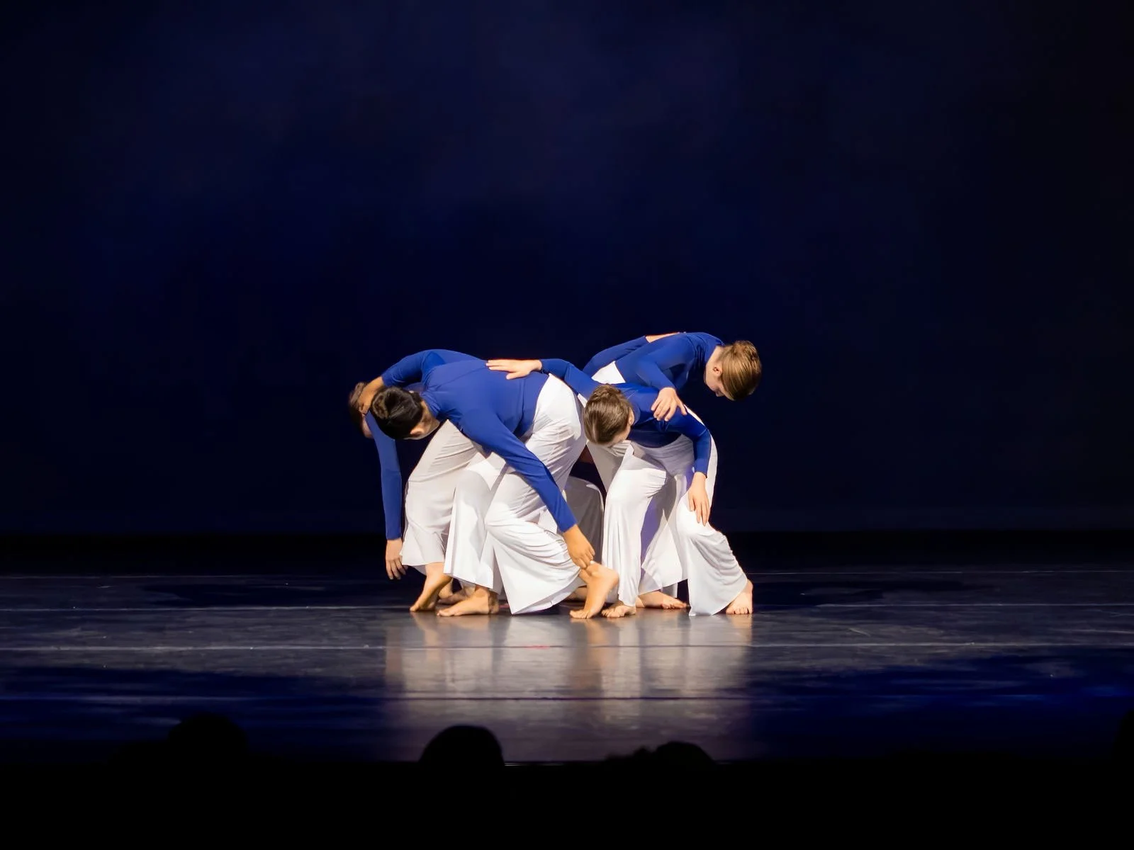 Four female dancers in blue tops and white pants performing a contemporary dance on stage, crouched close together, with a dark background.