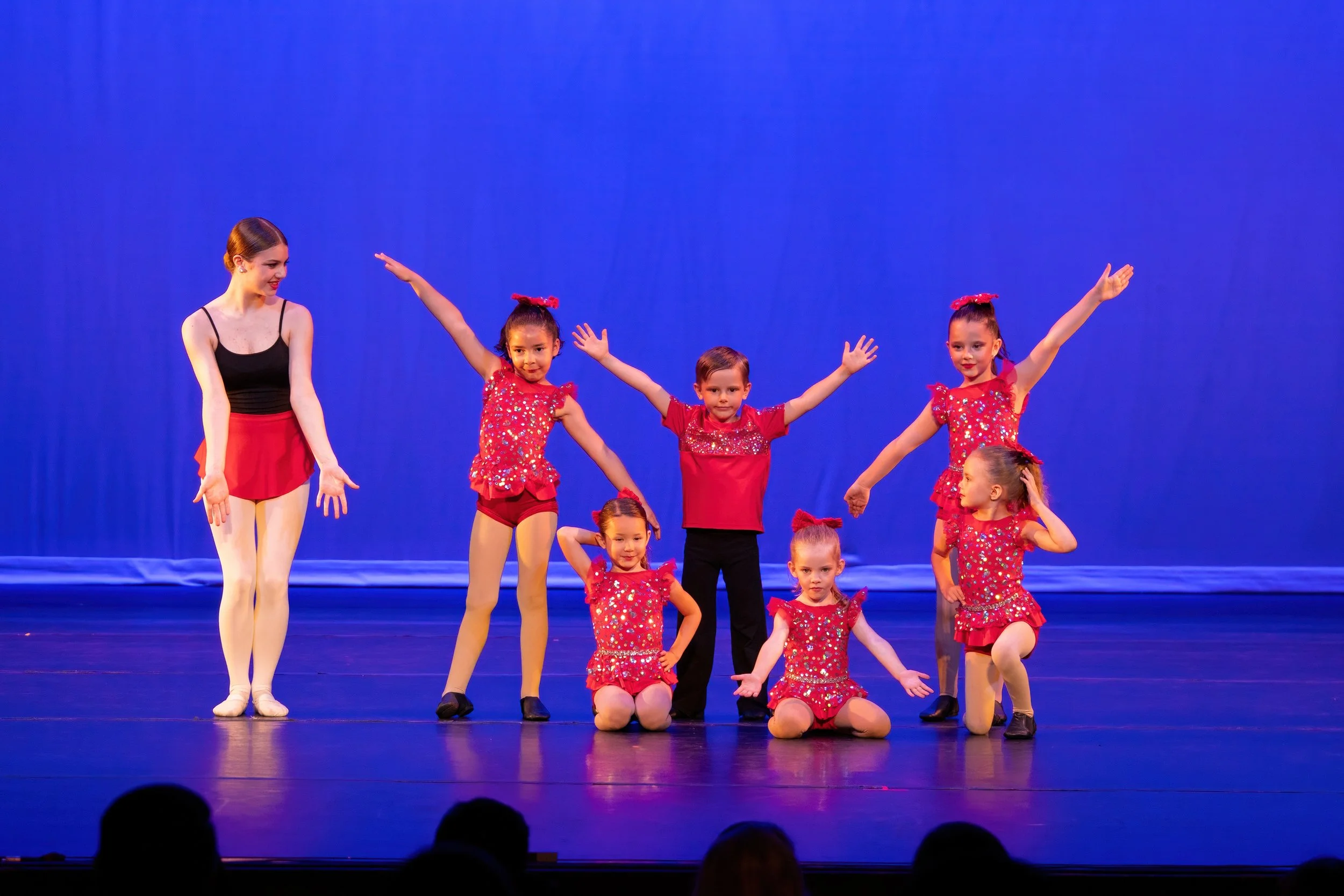 Group of young girls and a woman on stage during a dance recital, with the girls wearing red costumes with glitter, performing poses, and the woman in a black top with red skirt, in front of a blue curtain.