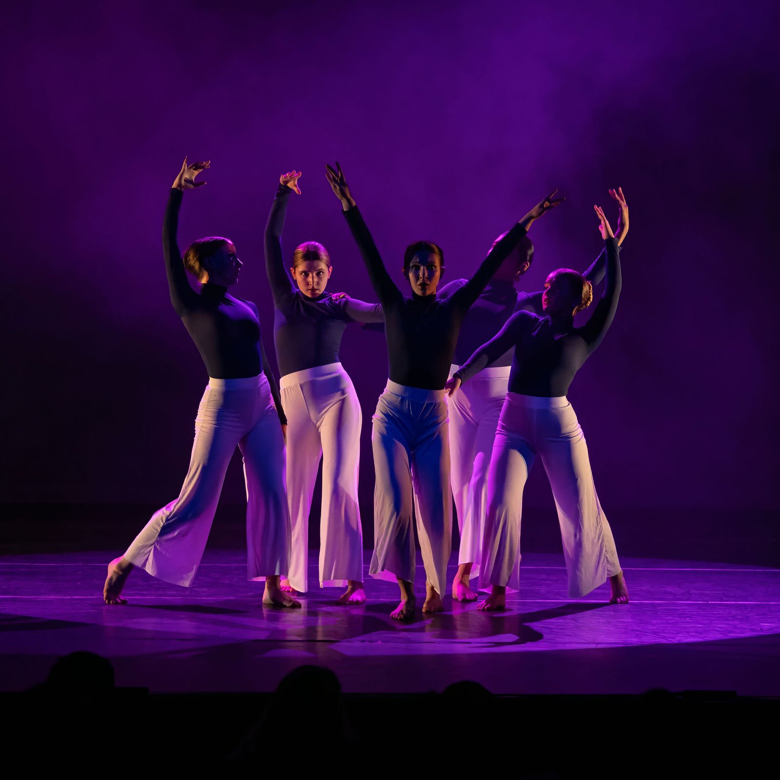 Five female dancers in black tops and white wide-leg pants perform on stage with purple lighting.