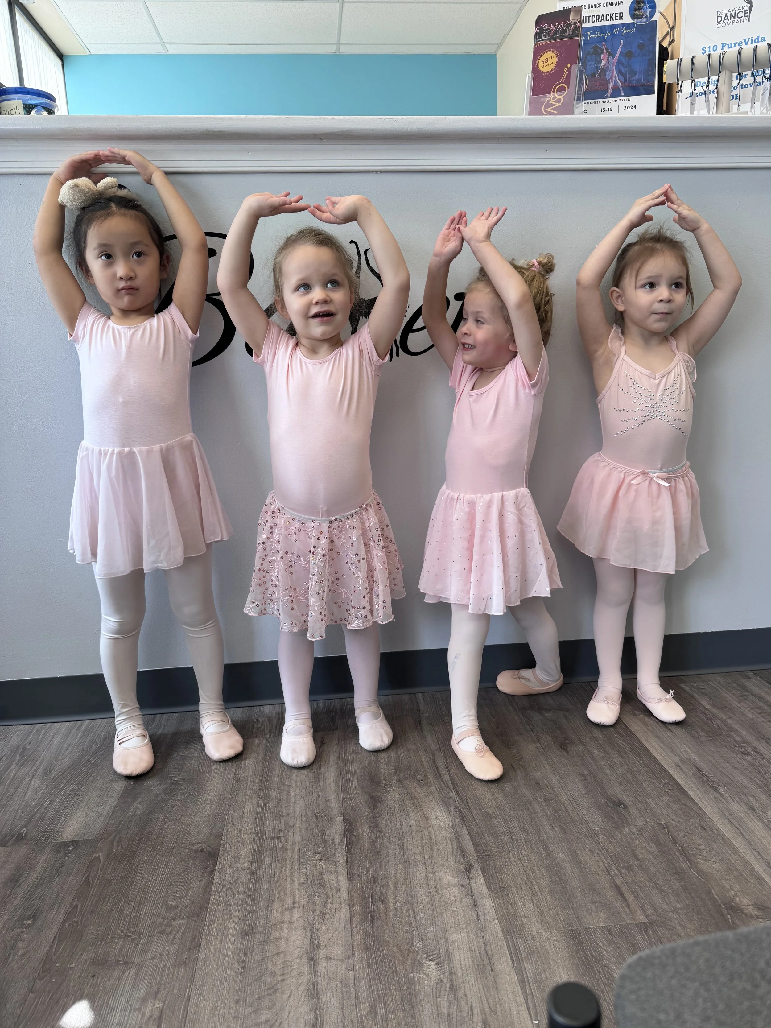 Four young girls in pink ballet dresses posing with their arms raised above their heads in a dance studio.