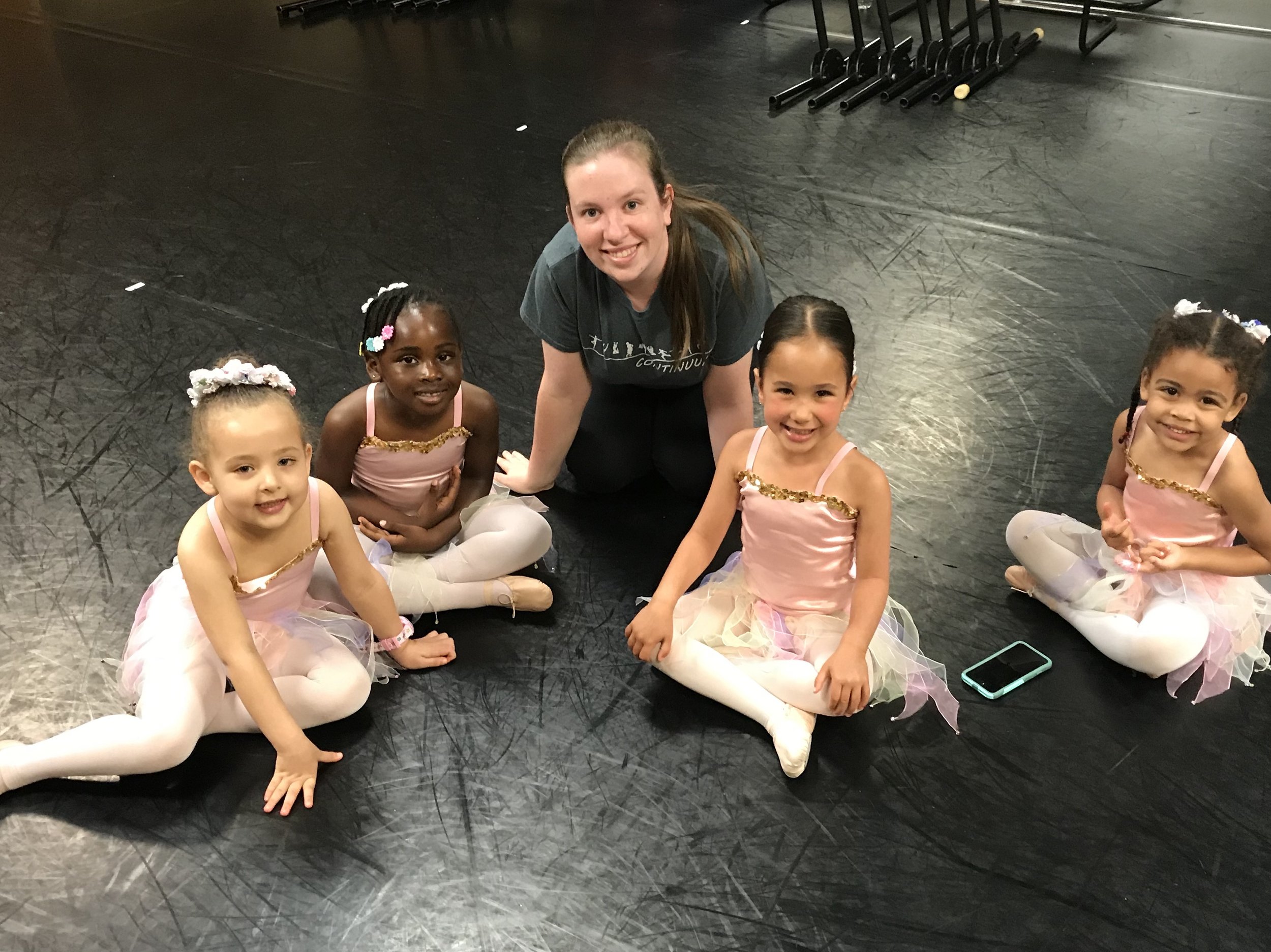 A young woman in casual clothing sitting on the floor with four young girls dressed in pink ballet tutus and white tights, smiling in a dance studio. The studio has a black floor, some equipment in the background, and a smartphone on the floor.