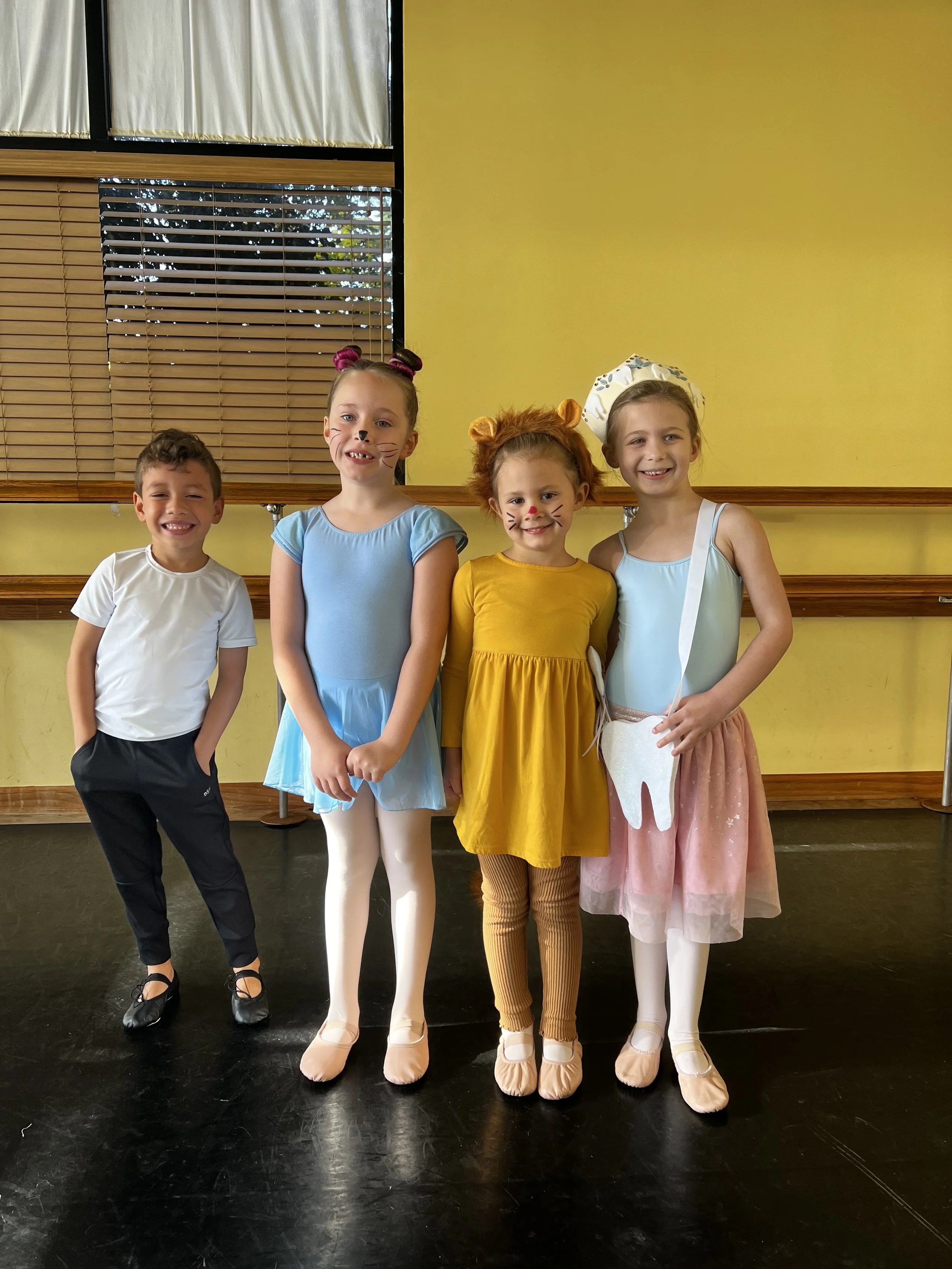 Four young children dressed in colorful costumes, with face paint, standing together in a dance studio with a yellow wall and wooden window blinds in the background.