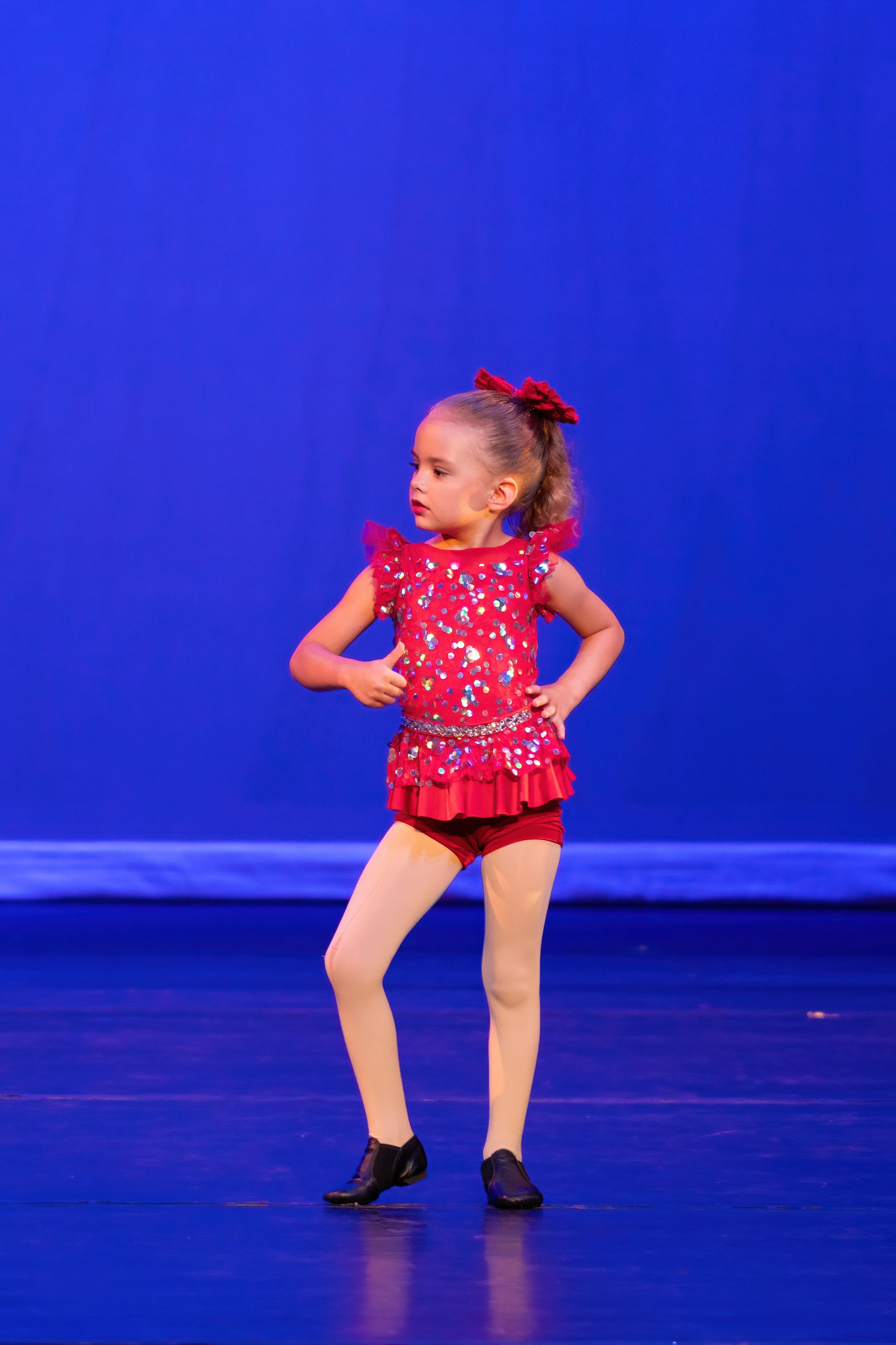 Young girl in a red sequined costume performing a dance on stage with a blue background.