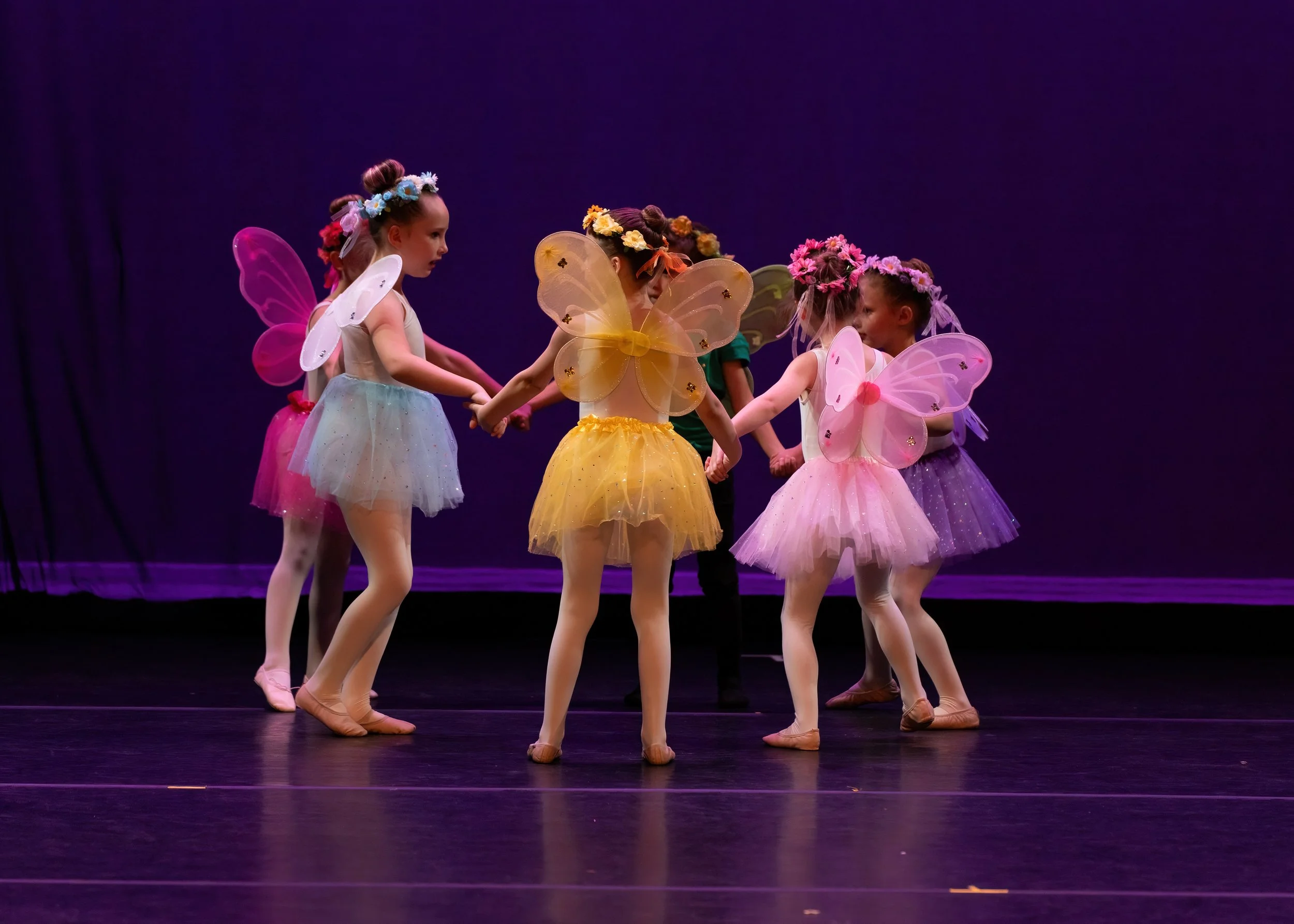 A group of young girls dressed as fairies holding hands in a ballet or dance performance on stage with a purple backdrop.