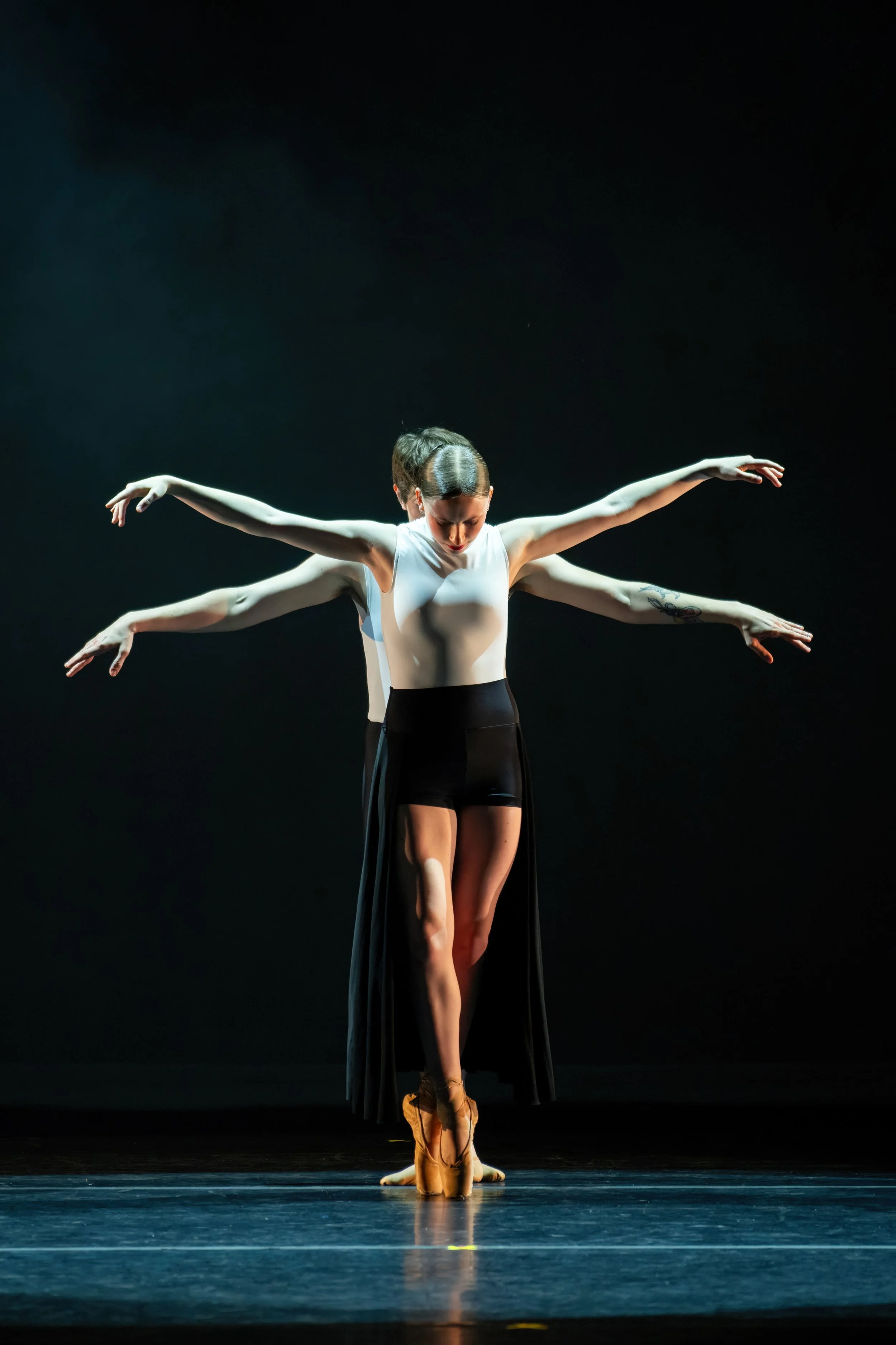 Ballet dancer on stage with advanced pose and three arms, wearing a white top, black shorts, and brown pointe shoes.