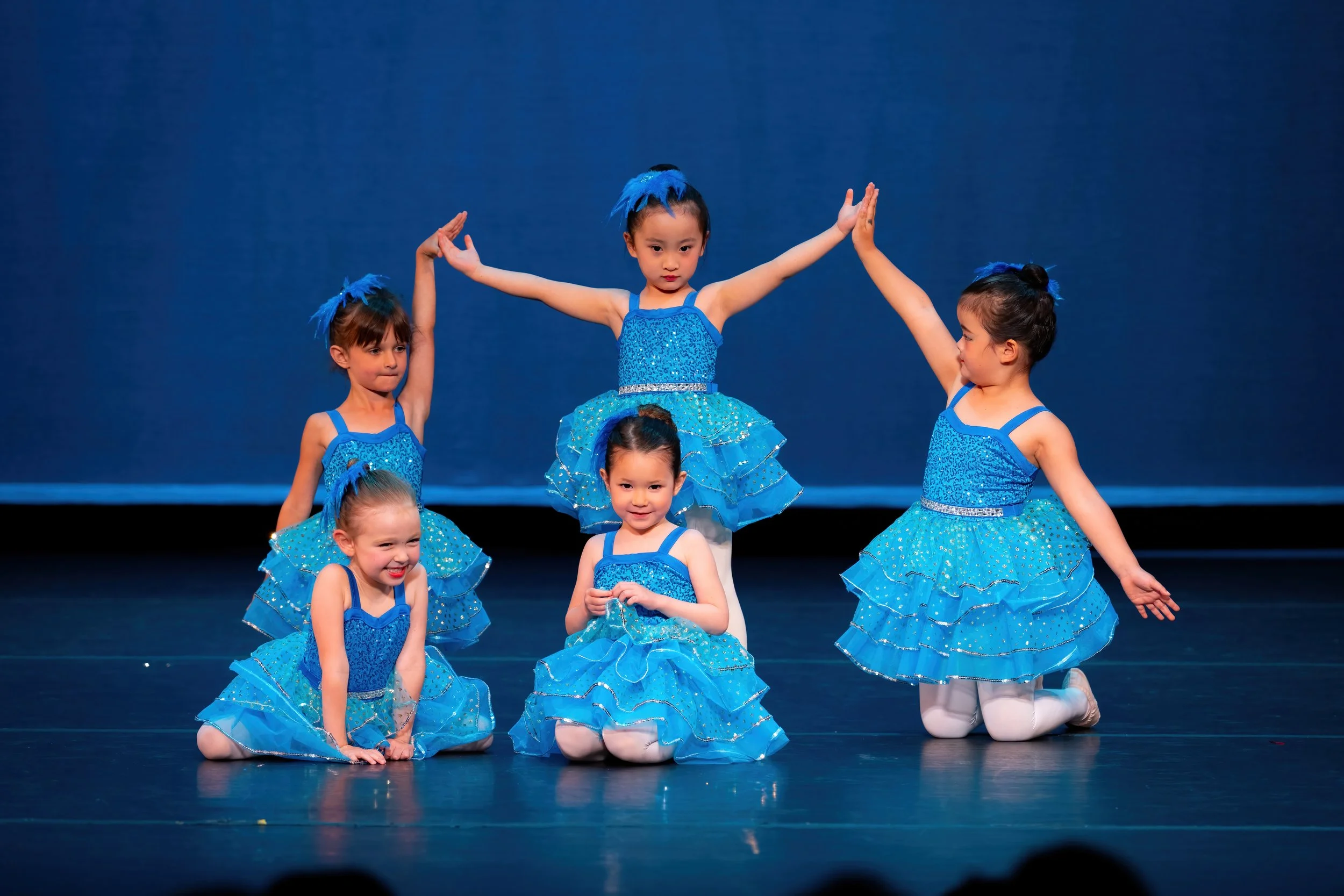 Five young girls in blue ballet costumes performing a dance on stage, with the girl in the center kneeling and smiling, and the others striking various poses.