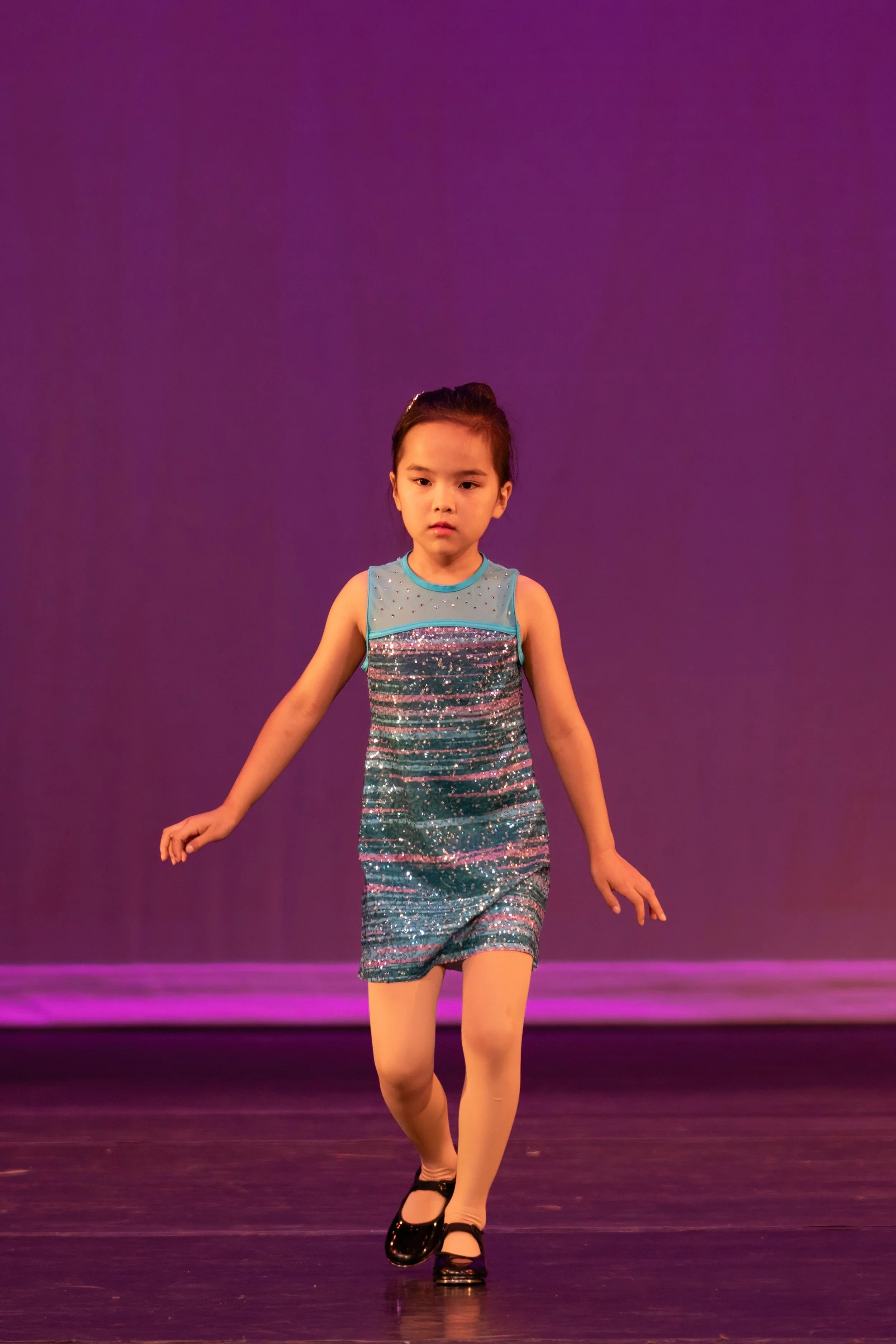 A young girl in a sparkly blue dress performing a dance on stage, with a purple background.