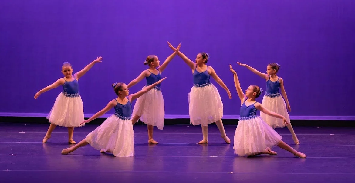 Six young ballerinas in blue and pink tutus performing a dance on stage with purple lighting.