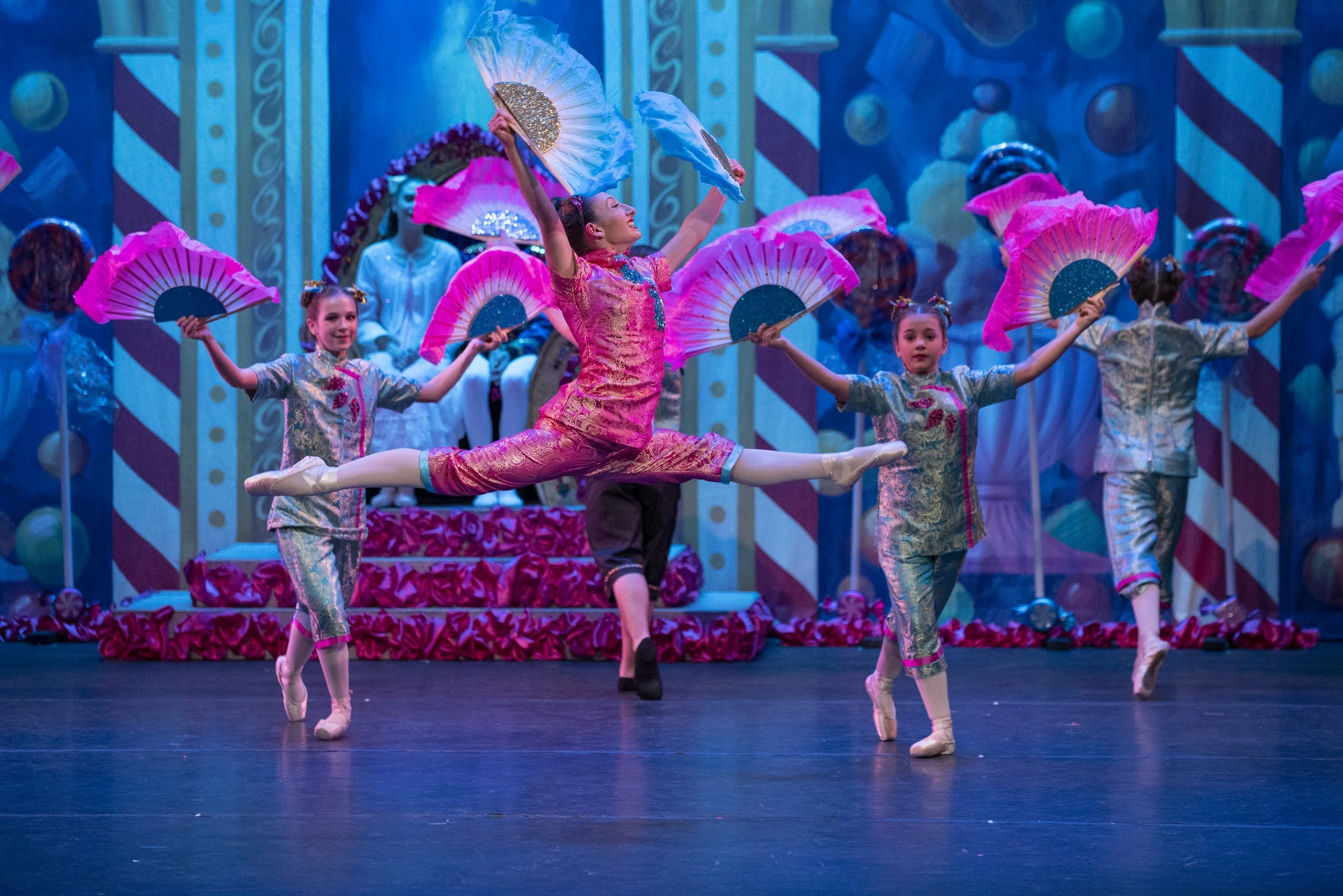 Children's ballet performance with dancers in colorful costumes holding pink and blue fans, on stage with vibrant backdrop and props.