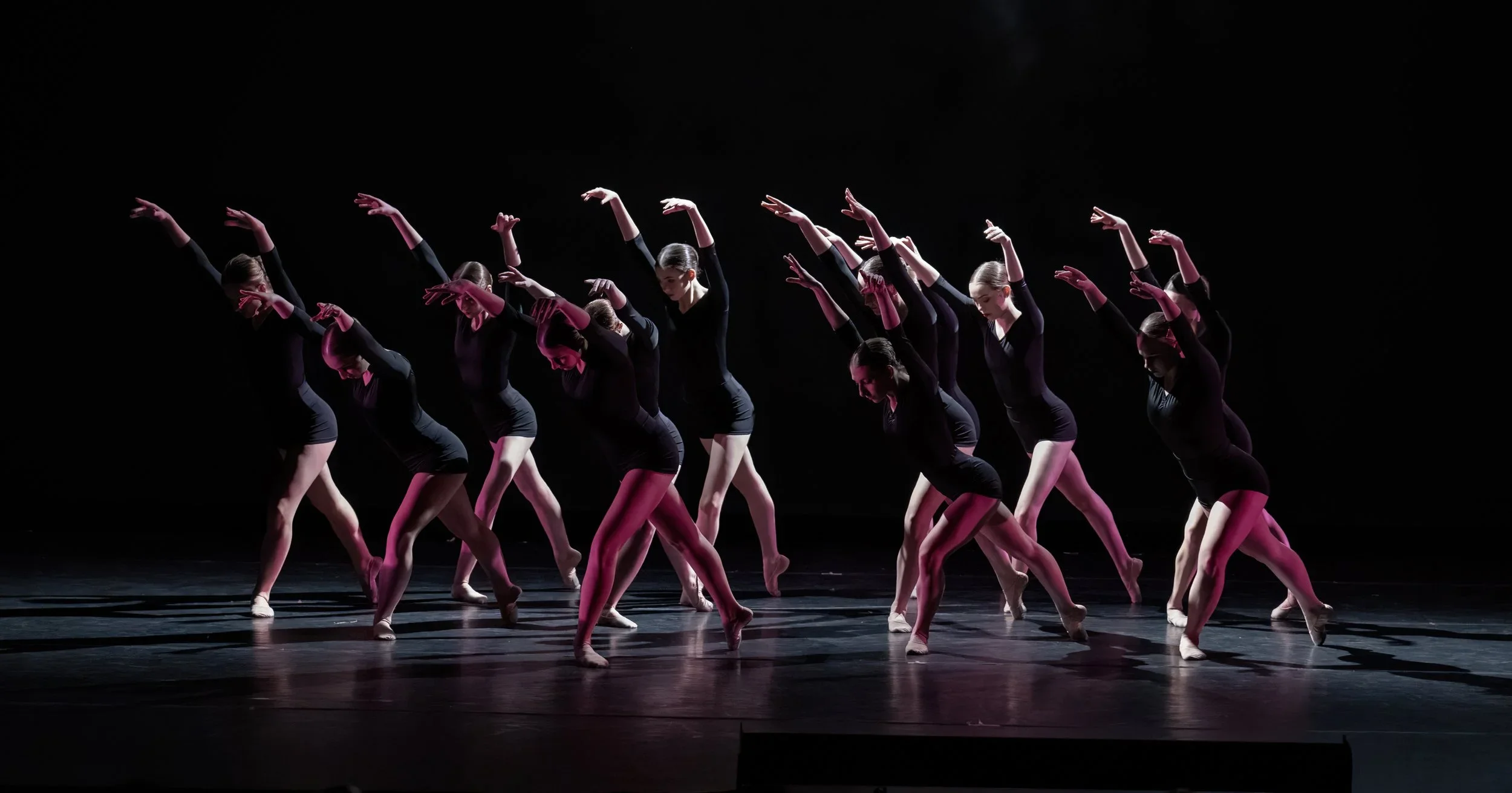 Group of female dancers in black costumes performing ballet on stage with dark background.